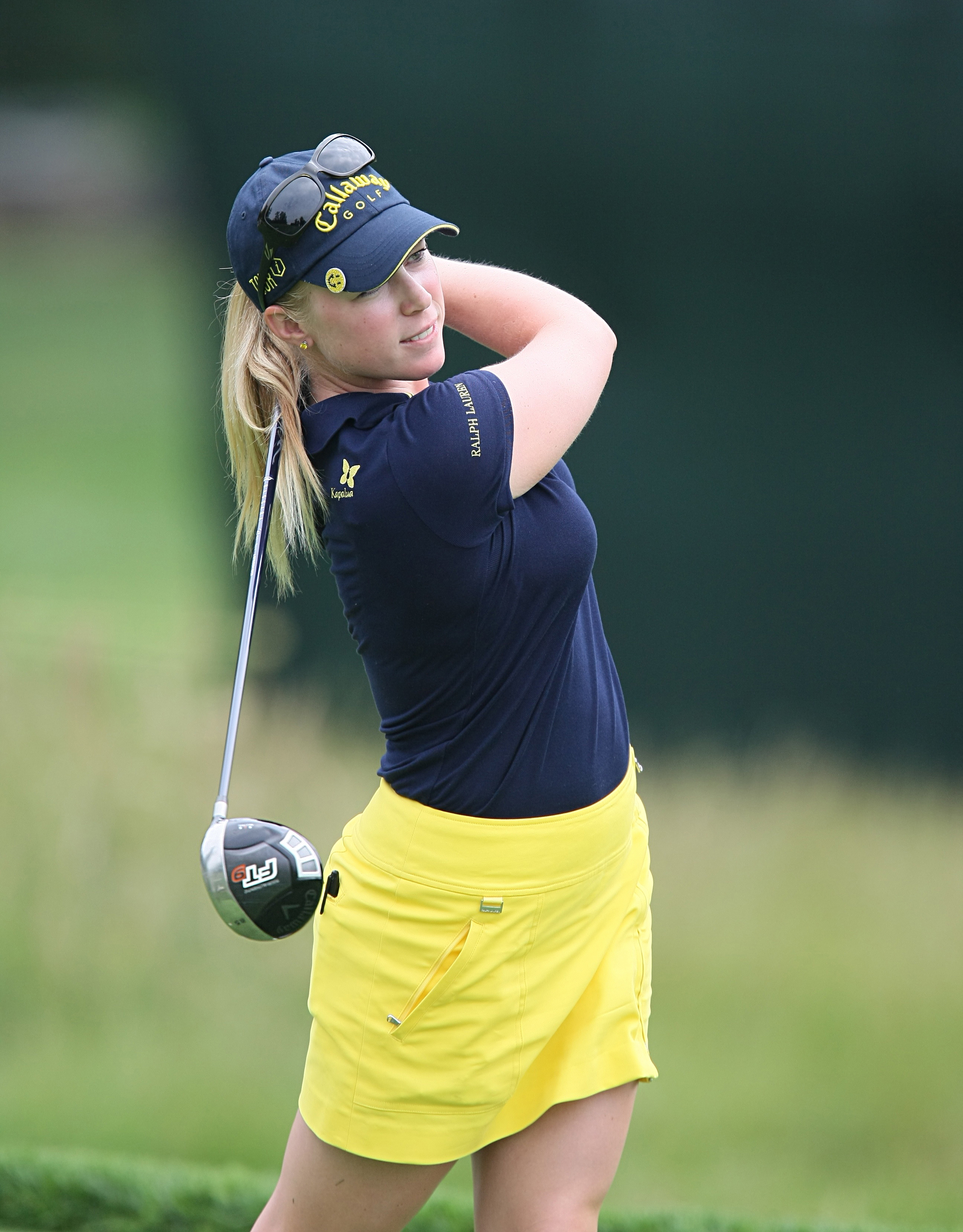 Female golfer in navy cap and polo shirt mid-swing with a driver on a golf course.