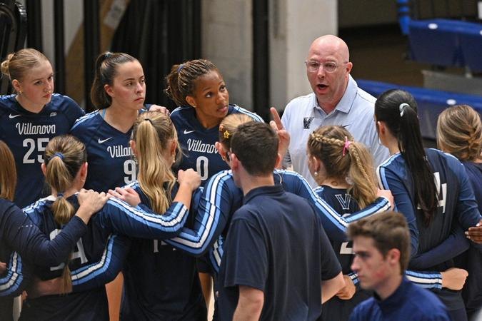 Volleyball players in Villanova uniforms huddle with a coach on an indoor court.