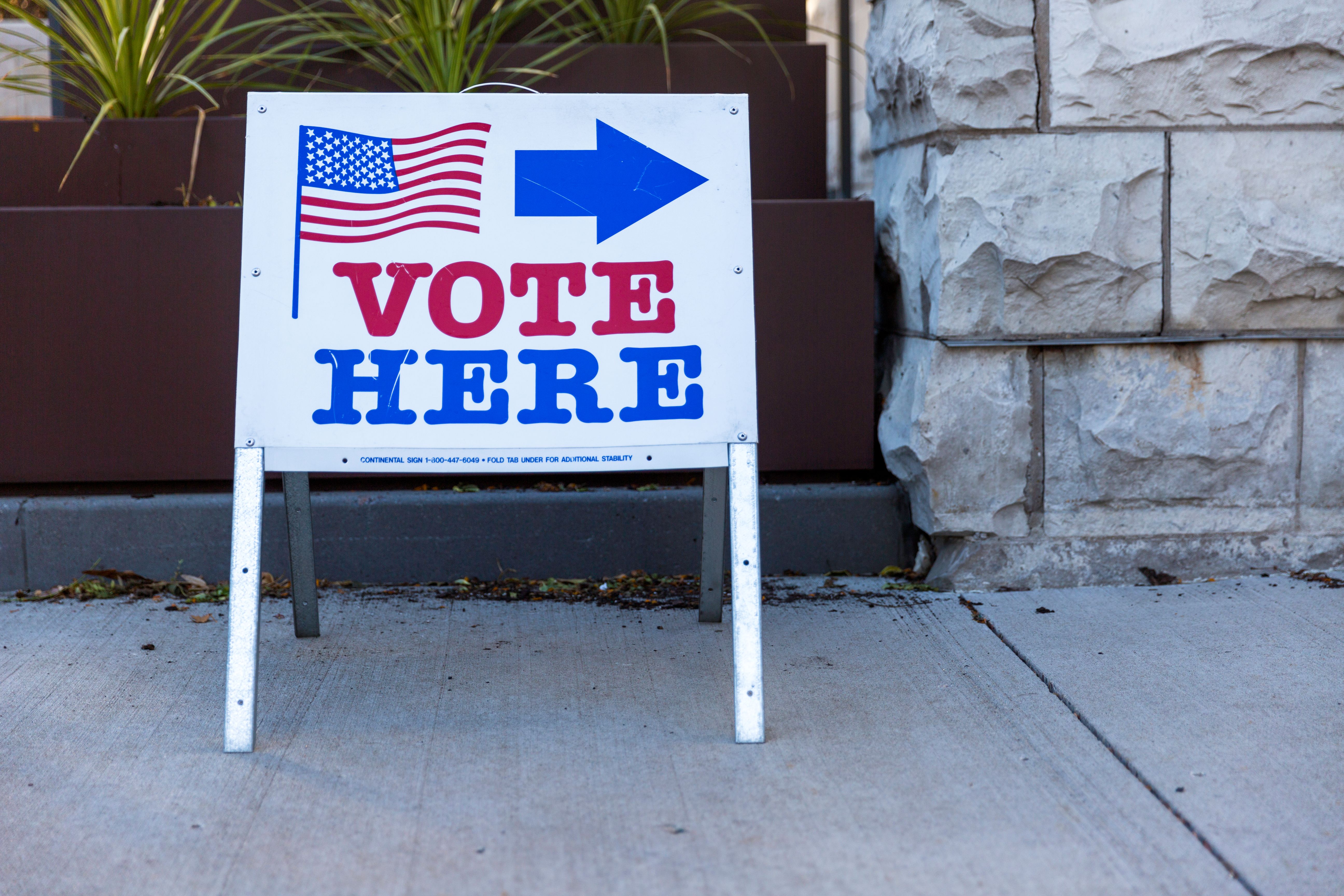A white 'Vote Here' A-frame sign with an American flag on a sidewalk outside a polling place.