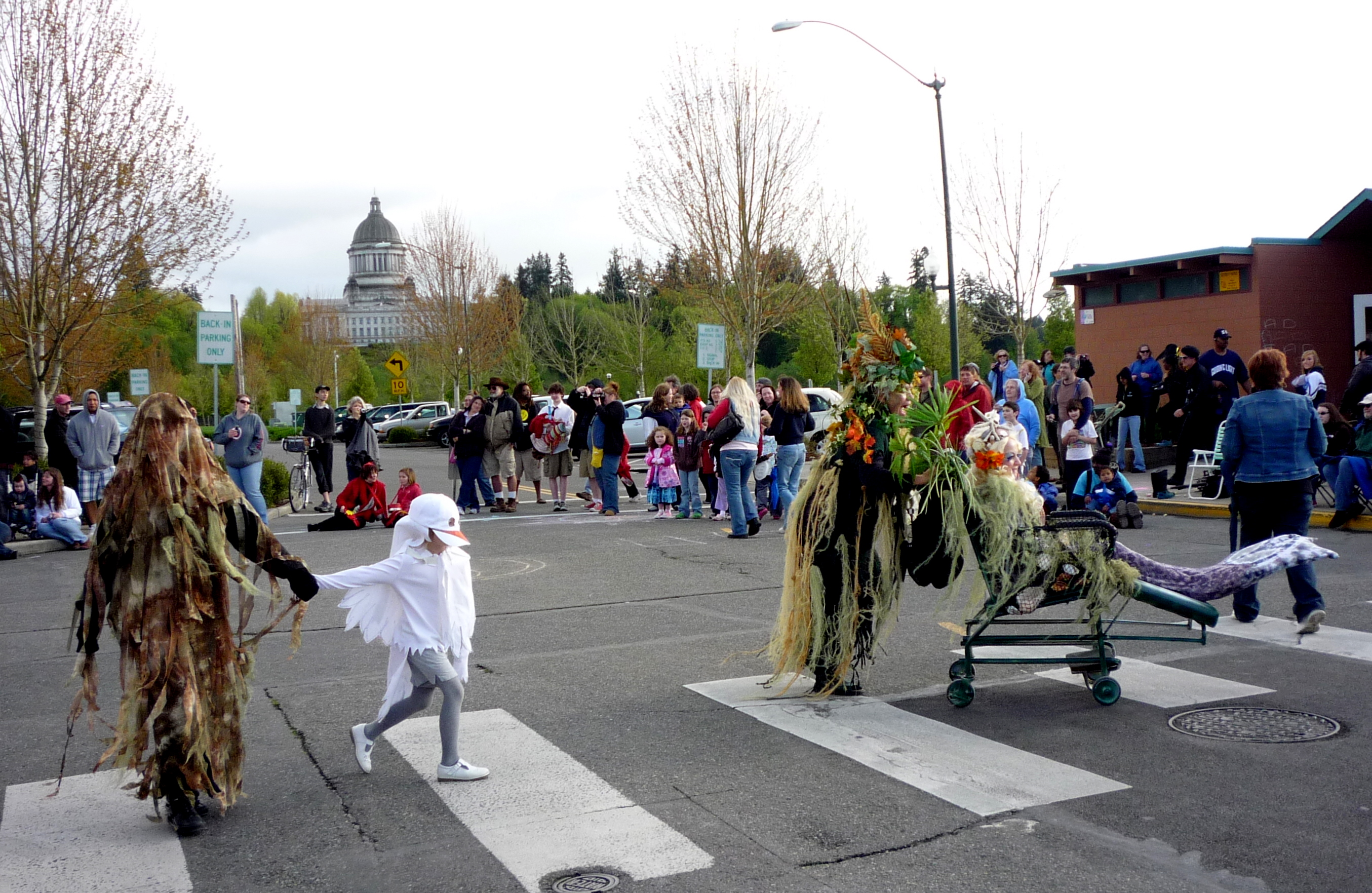 Costumed parade performers cross a crosswalk with a domed capitol building visible in the background.