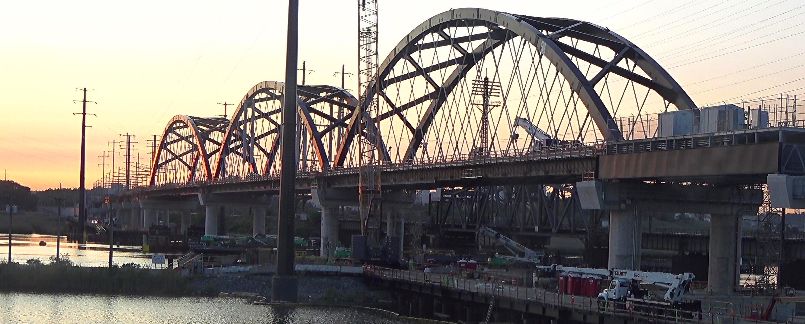 Steel arch railroad bridge spanning a wide river at dusk.
