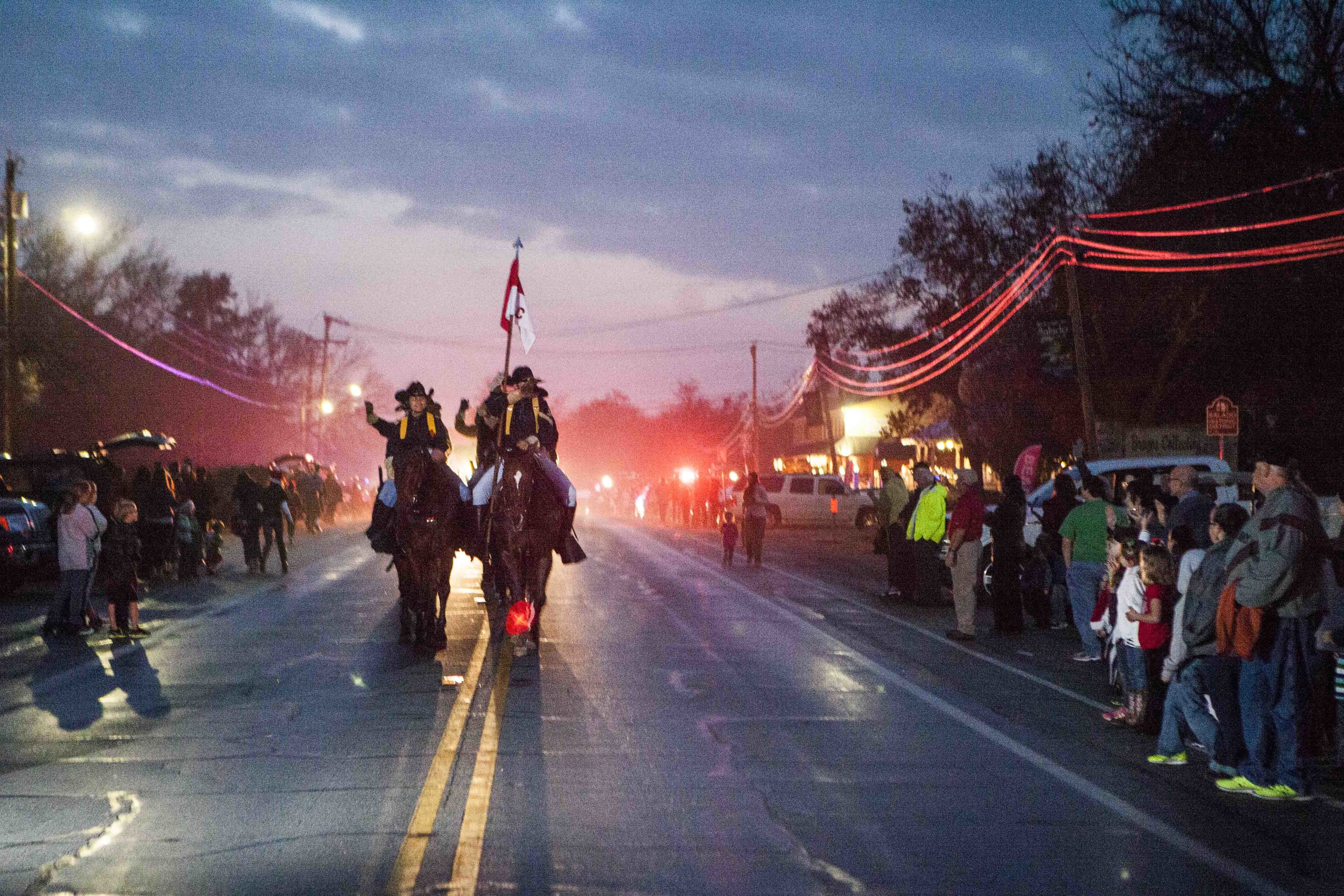 Two riders on horseback lead a Christmas parade down a street at dusk with crowds on both sides.