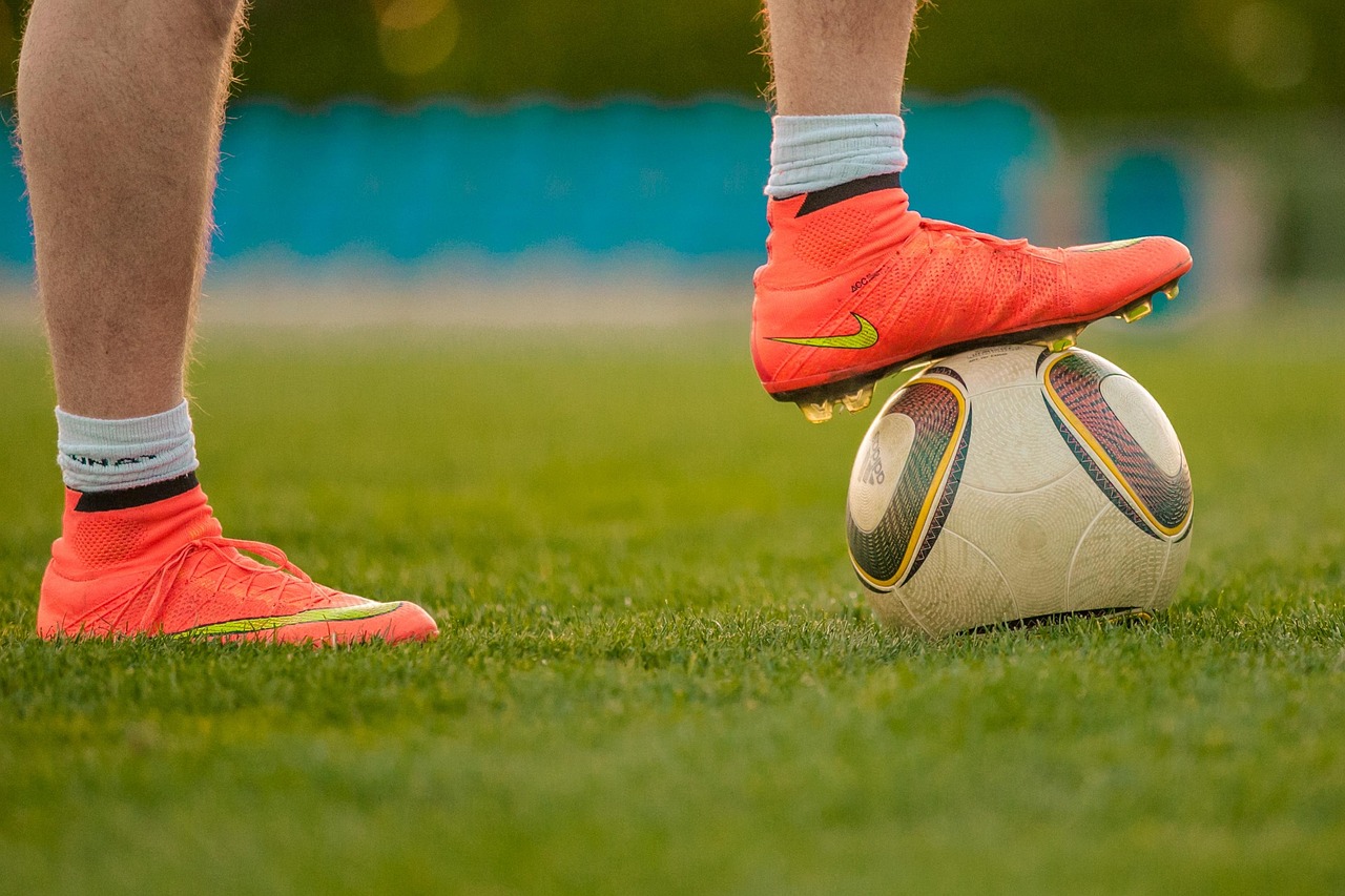 Close-up of a soccer player's legs, wearing orange cleats with one foot resting on top of the ball.