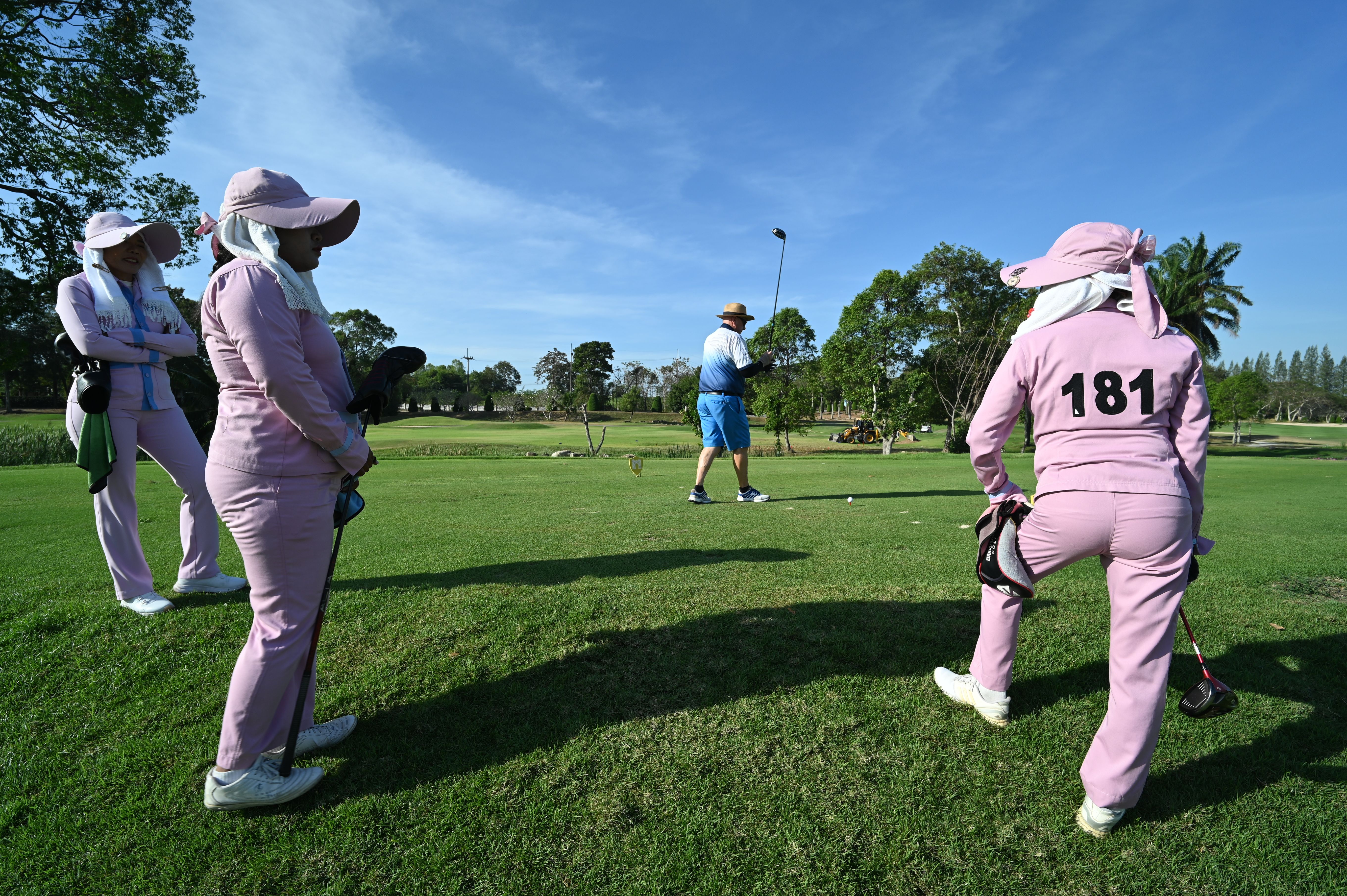 Several women golfers in matching uniforms watch a teammate swing on a sunny course.