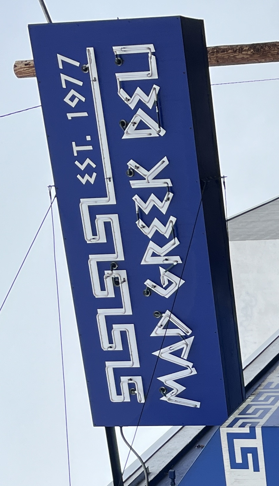 Blue illuminated sign reading Mad Greek Deli Est. 1977 with decorative Greek border