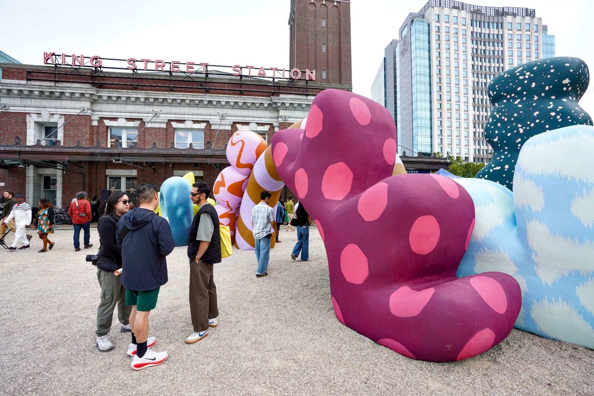 Large colorful polka-dotted sculpture on a plaza with visitors walking around it and city buildings in the background.