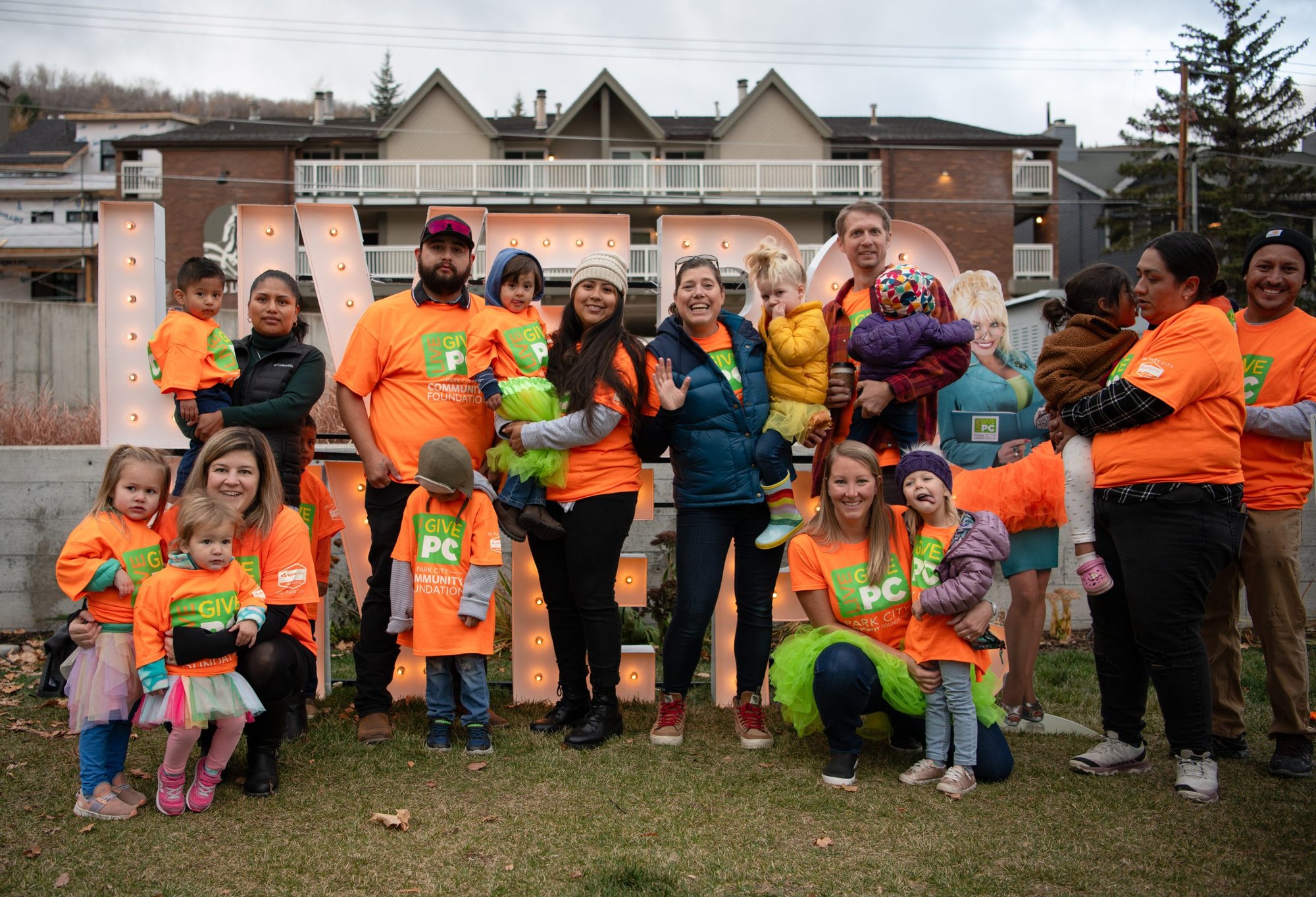 Group of adults and children in orange T-shirts posing outdoors in front of illuminated marquee letters.
