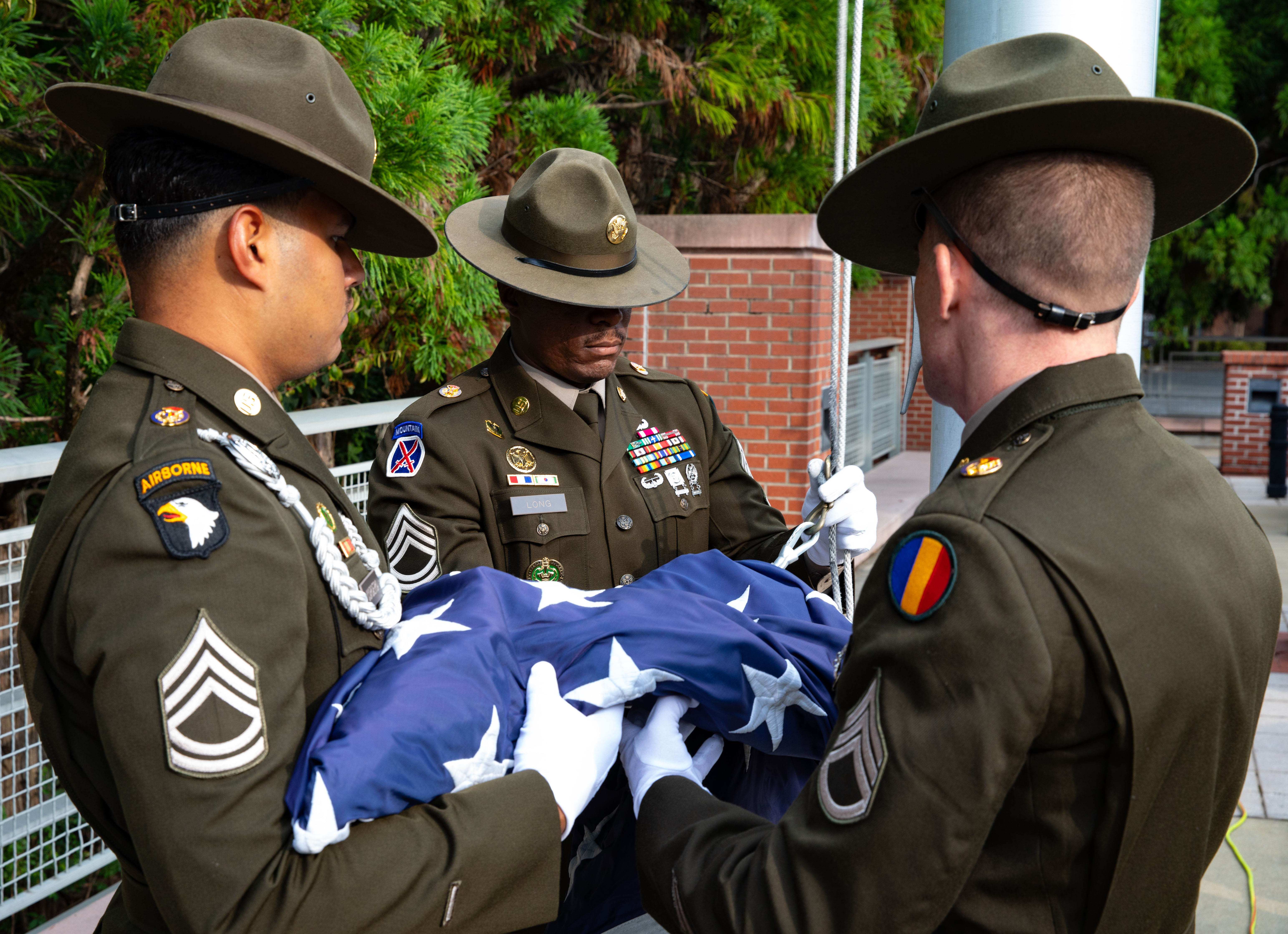 Three soldiers in dress uniforms and white gloves unfurl an American flag outdoors.