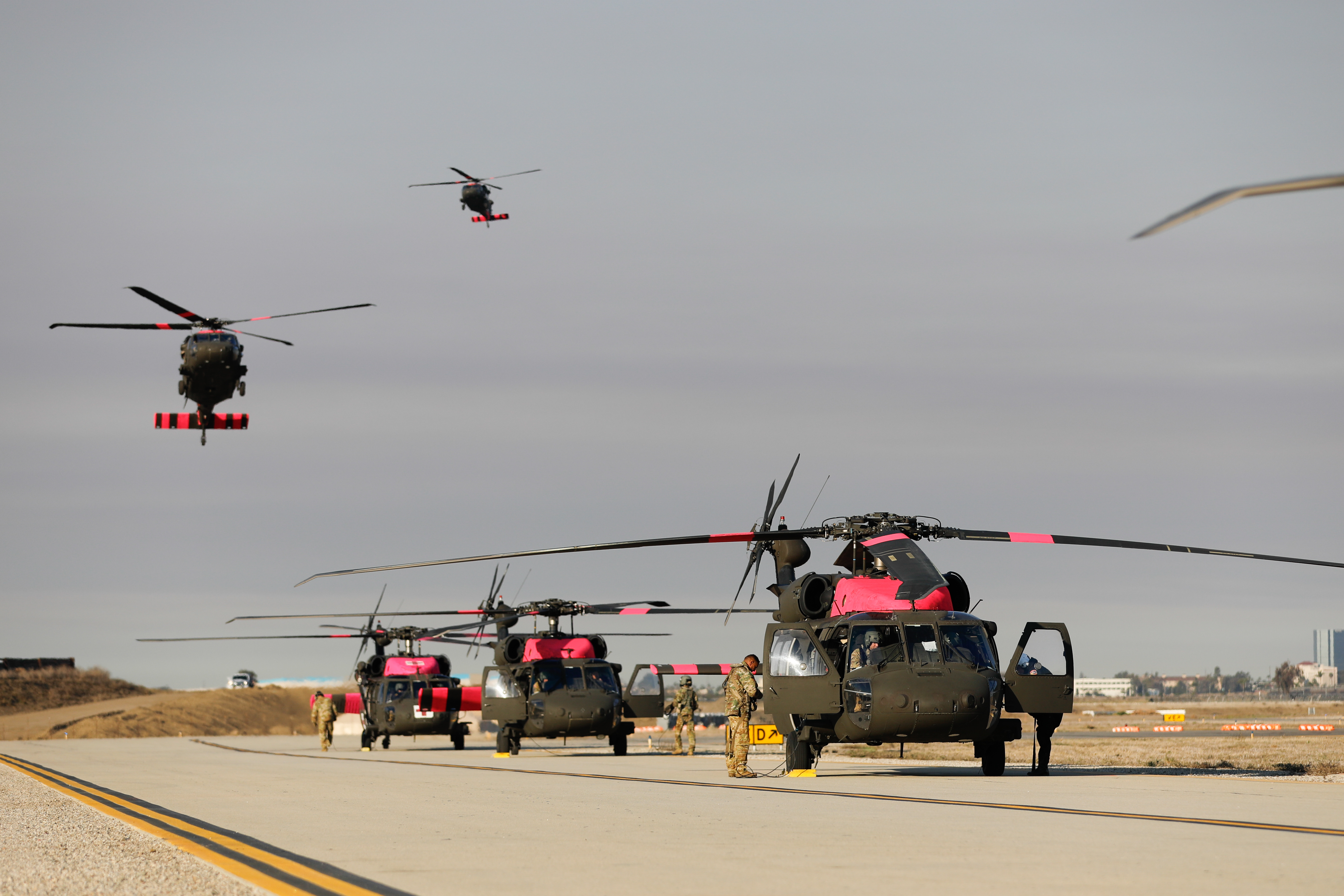 Military helicopters on an airport tarmac with two in flight above.
