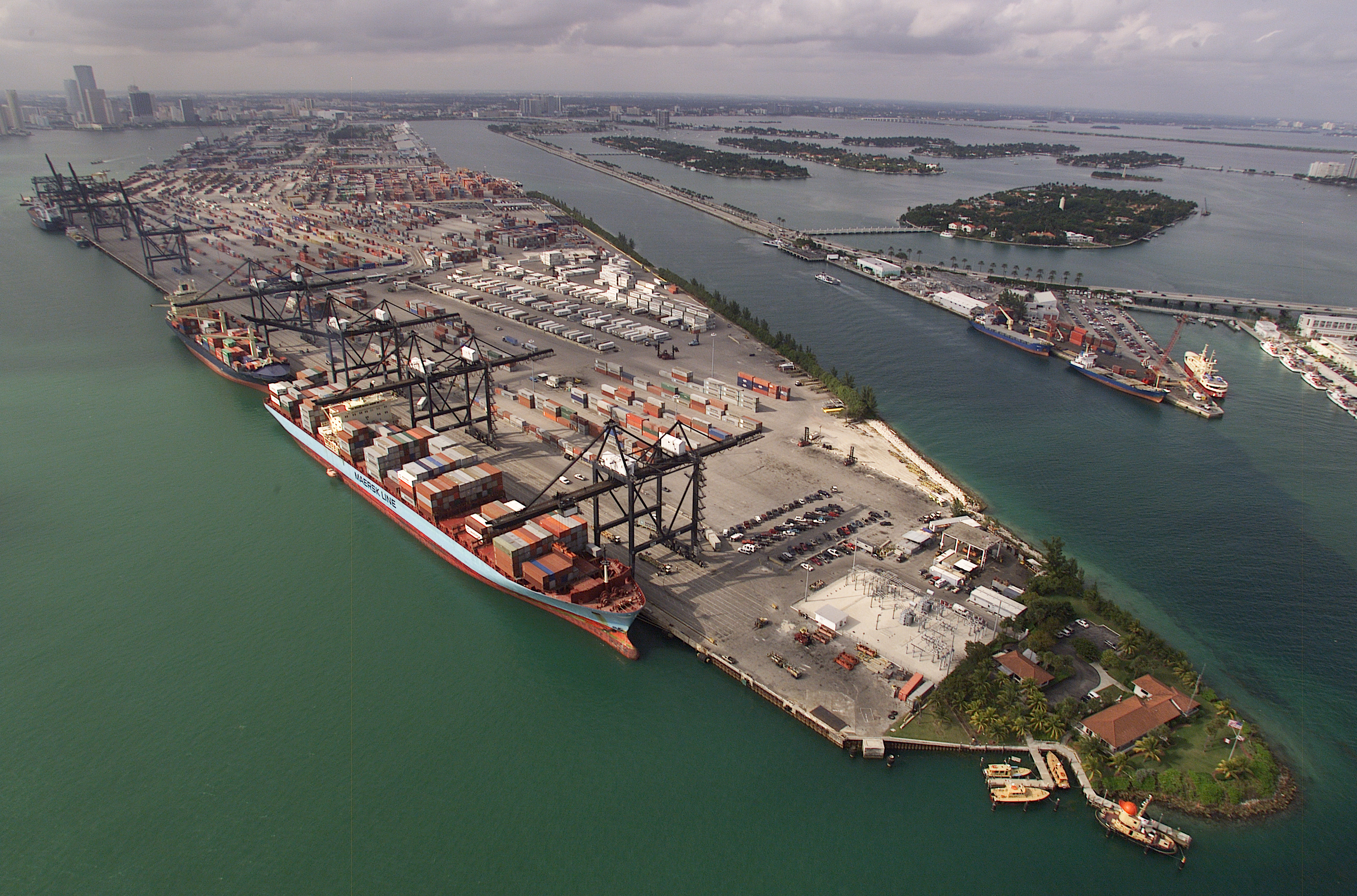 Aerial view of a large container port with cranes, stacked shipping containers, and a city skyline beyond.