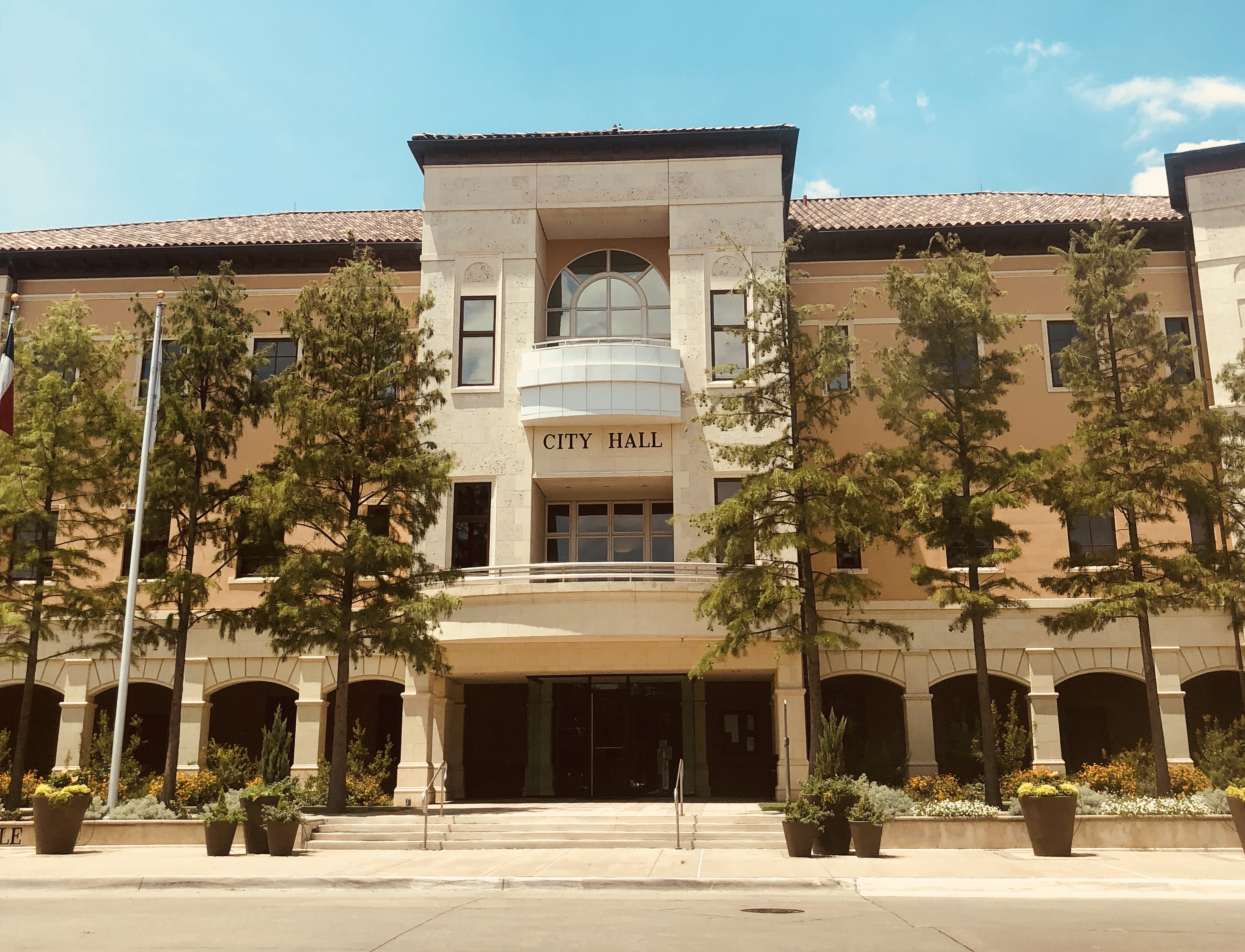 Two-story beige city hall building with arched entrance, trees, and flower planters.