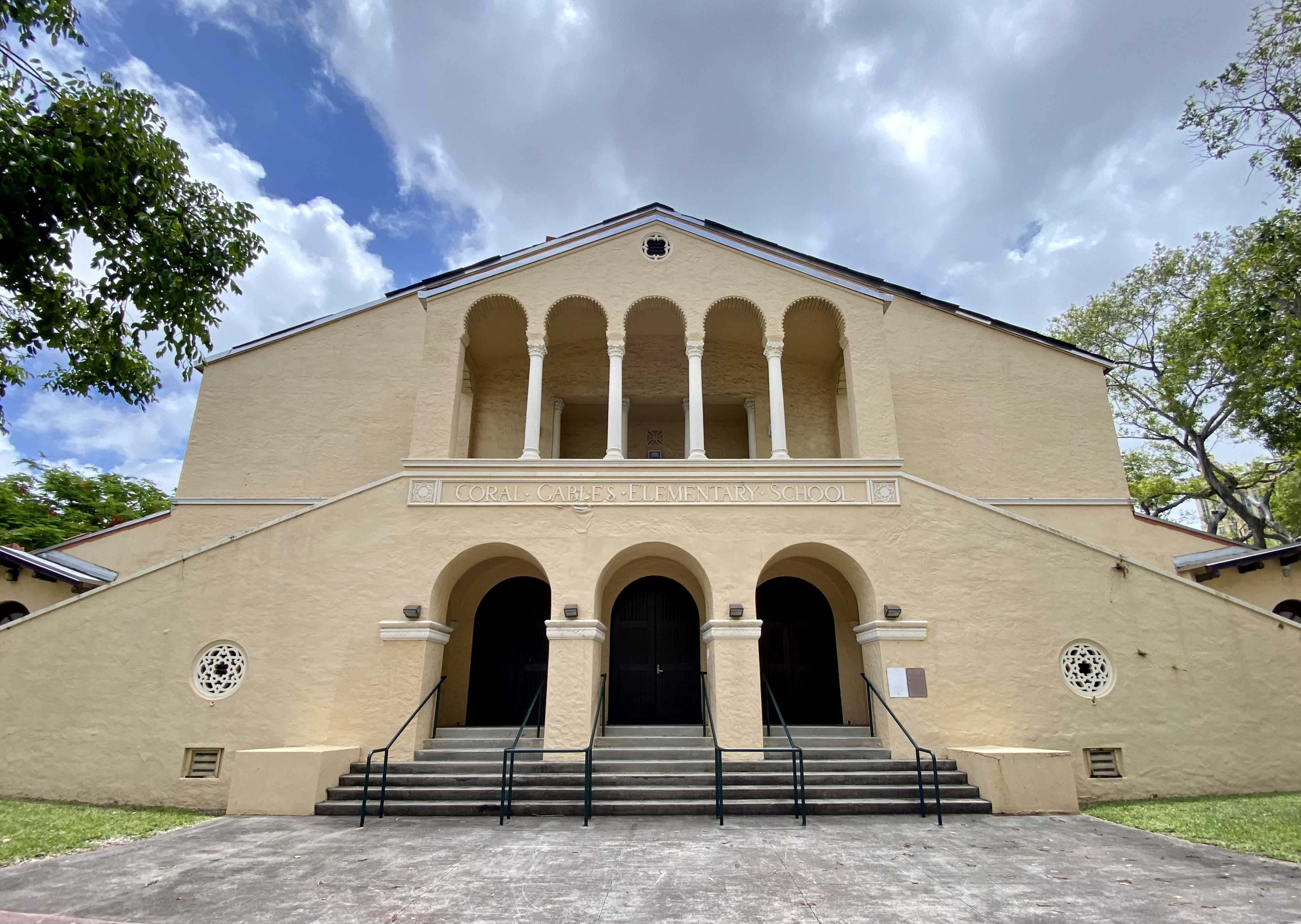 Yellow stucco school building with arched entryways and a colonnaded upper balcony.