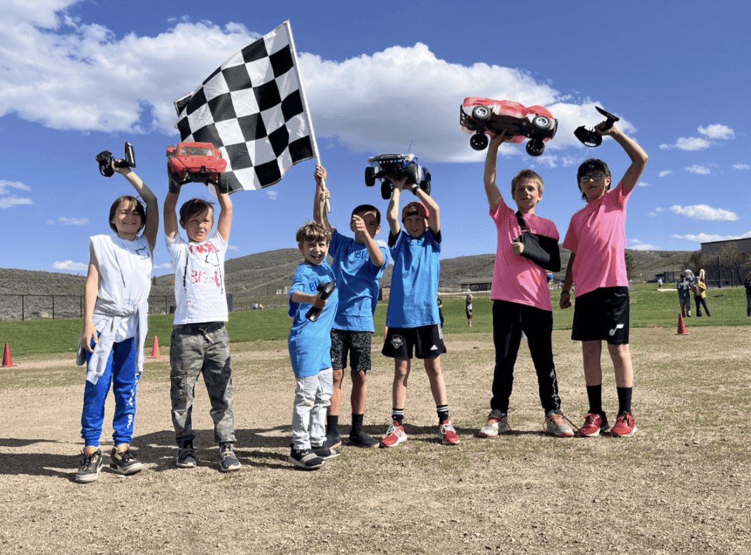 Children outdoors holding remote-control monster trucks and a checkered racing flag.