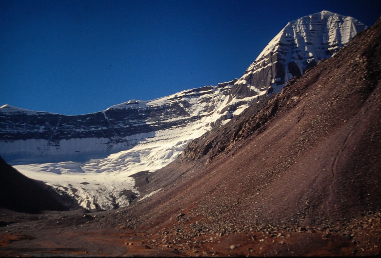 Snow-capped mountain with glaciers and a rocky scree slope under a deep blue sky.