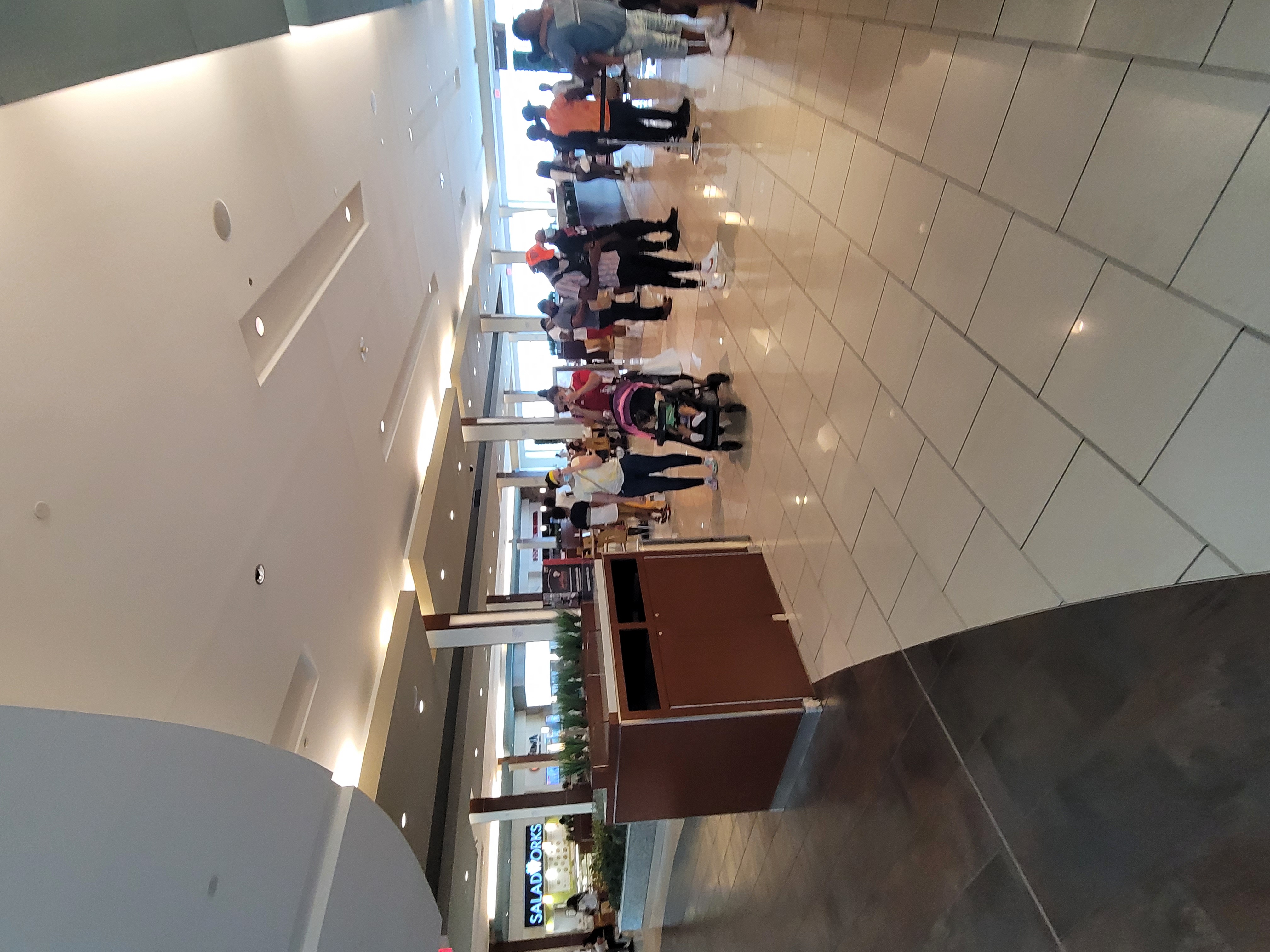 Shoppers walk through a shopping mall corridor past a central kiosk and retail storefronts.