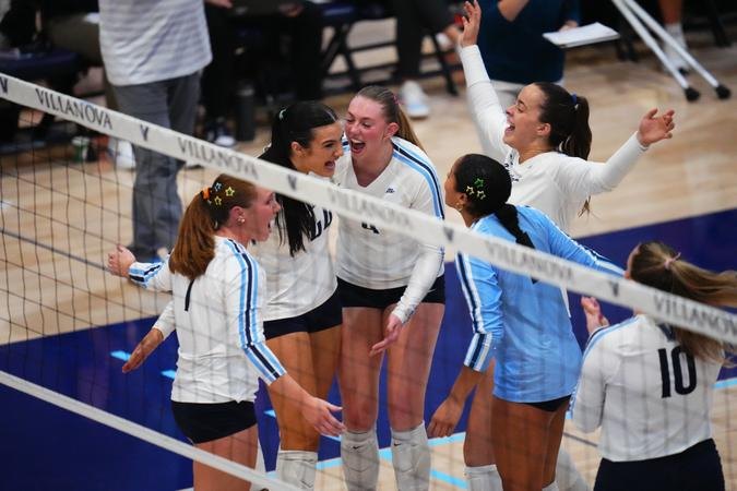 Female volleyball players in white and blue uniforms huddle near the net in celebration.