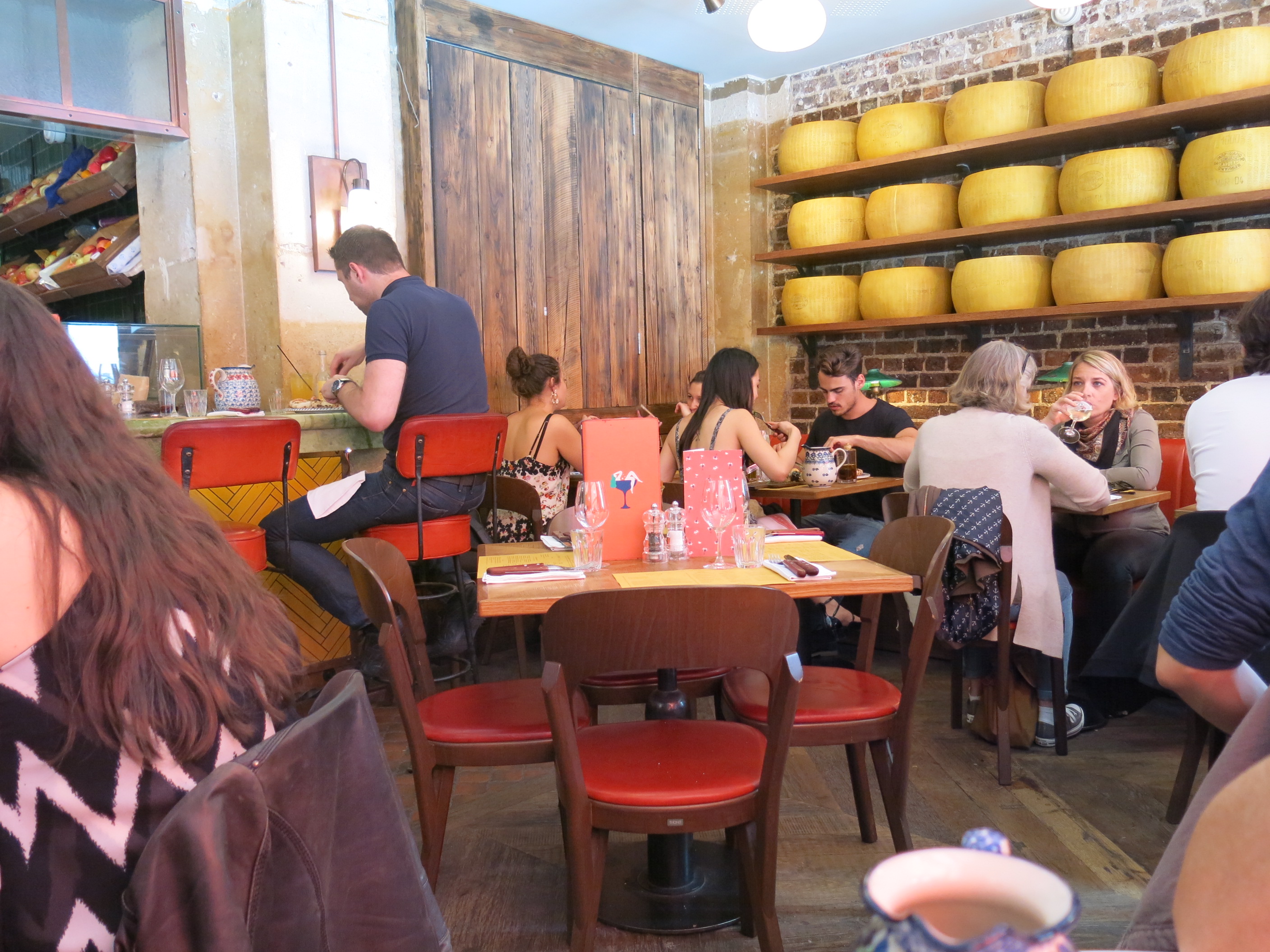 Crowded restaurant dining room with red chairs, wooden decor, and cheese wheels on shelves.