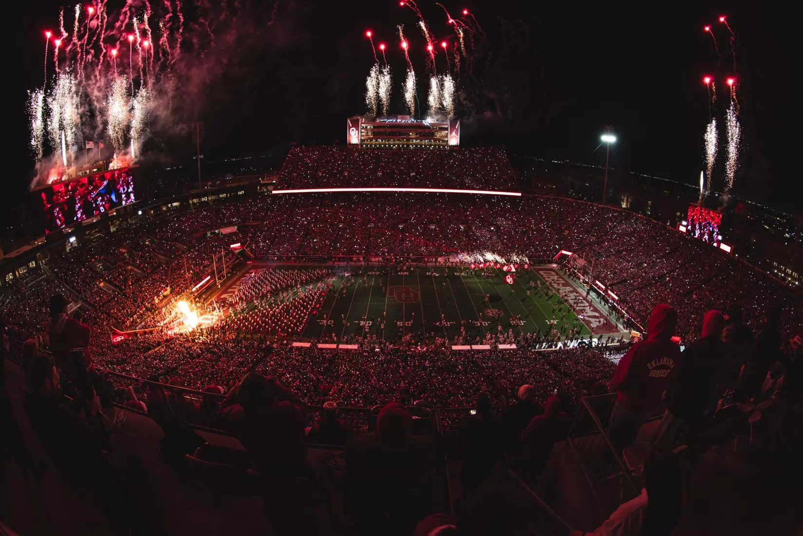 Fisheye aerial view of a packed football stadium at night with fireworks overhead.