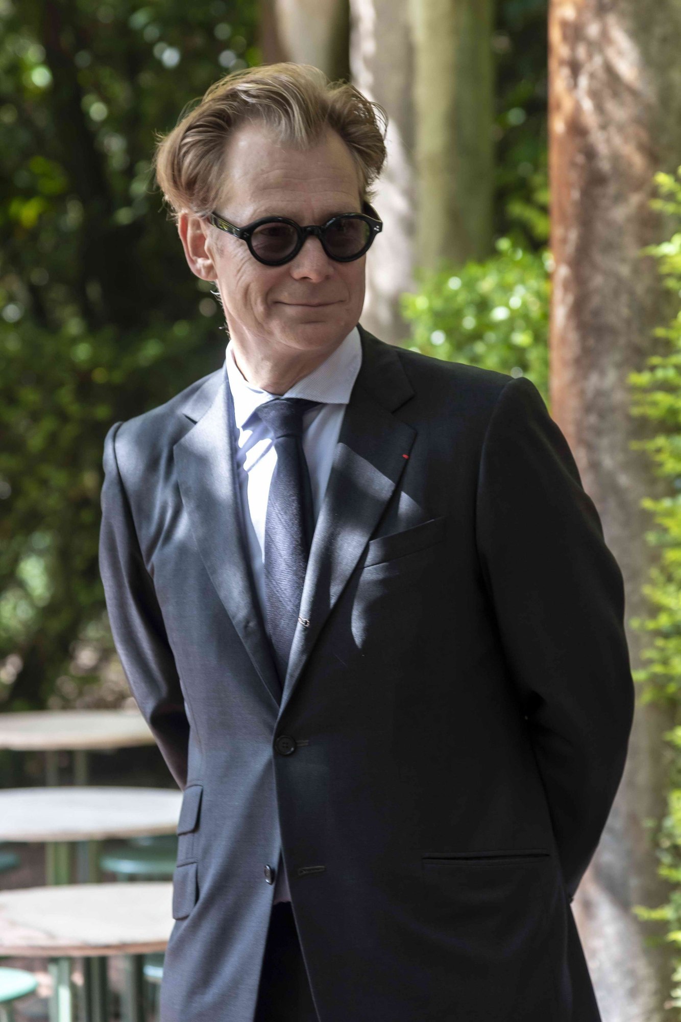 Man in dark suit and round sunglasses standing outdoors among trees.