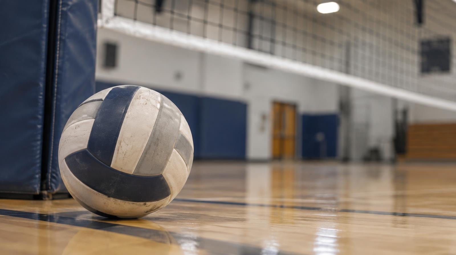 A volleyball resting on a hardwood gymnasium floor near a net post.