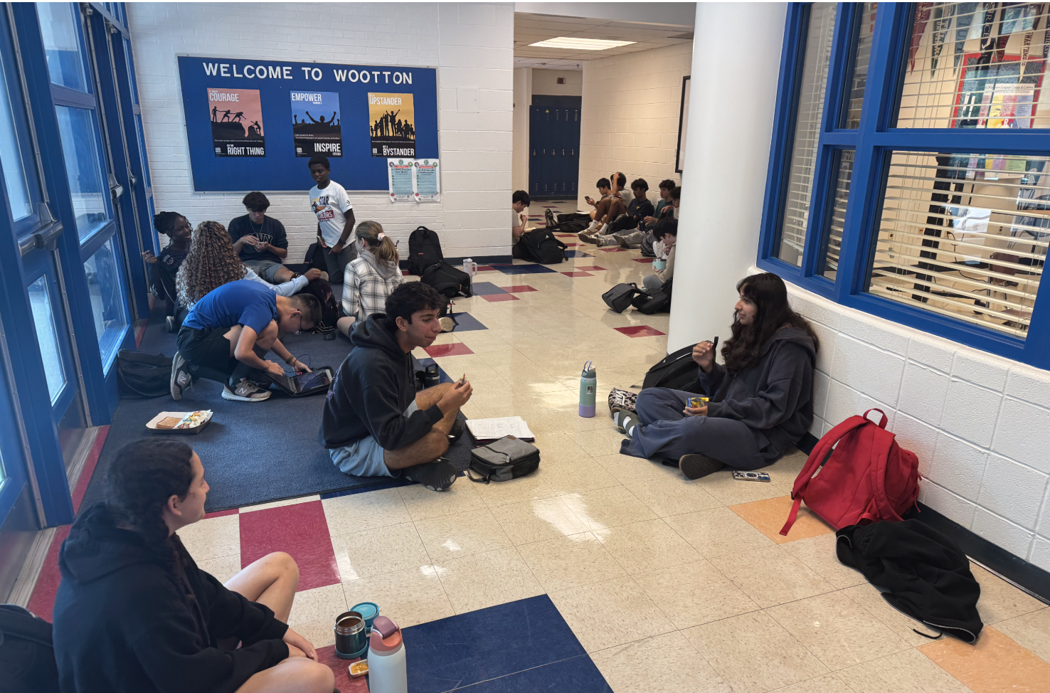 Students seated on a school hallway floor with backpacks and food, a welcome banner on the wall.