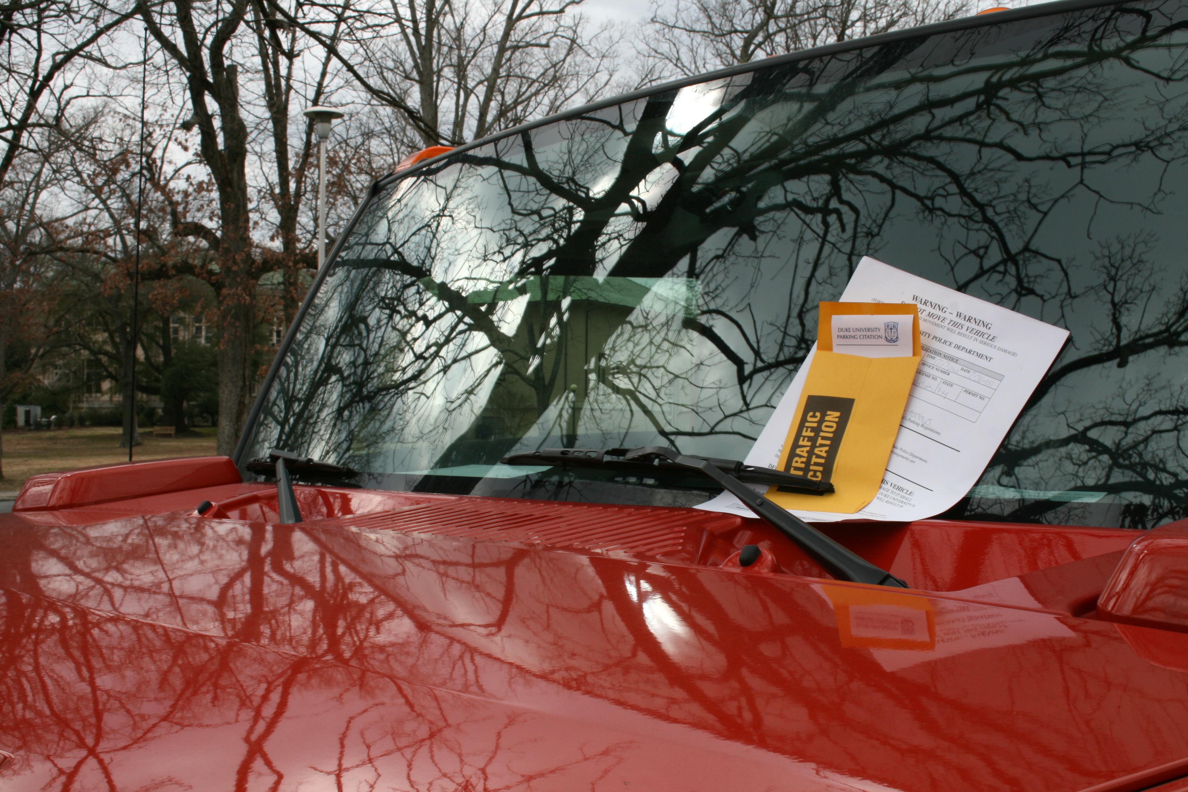 A yellow traffic citation envelope under the windshield wiper of a red SUV parked outdoors.