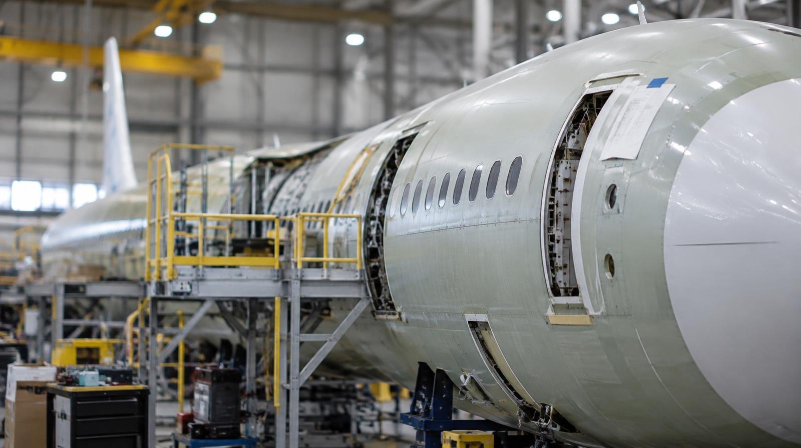 A large aircraft fuselage surrounded by scaffolding inside a bright industrial hangar.