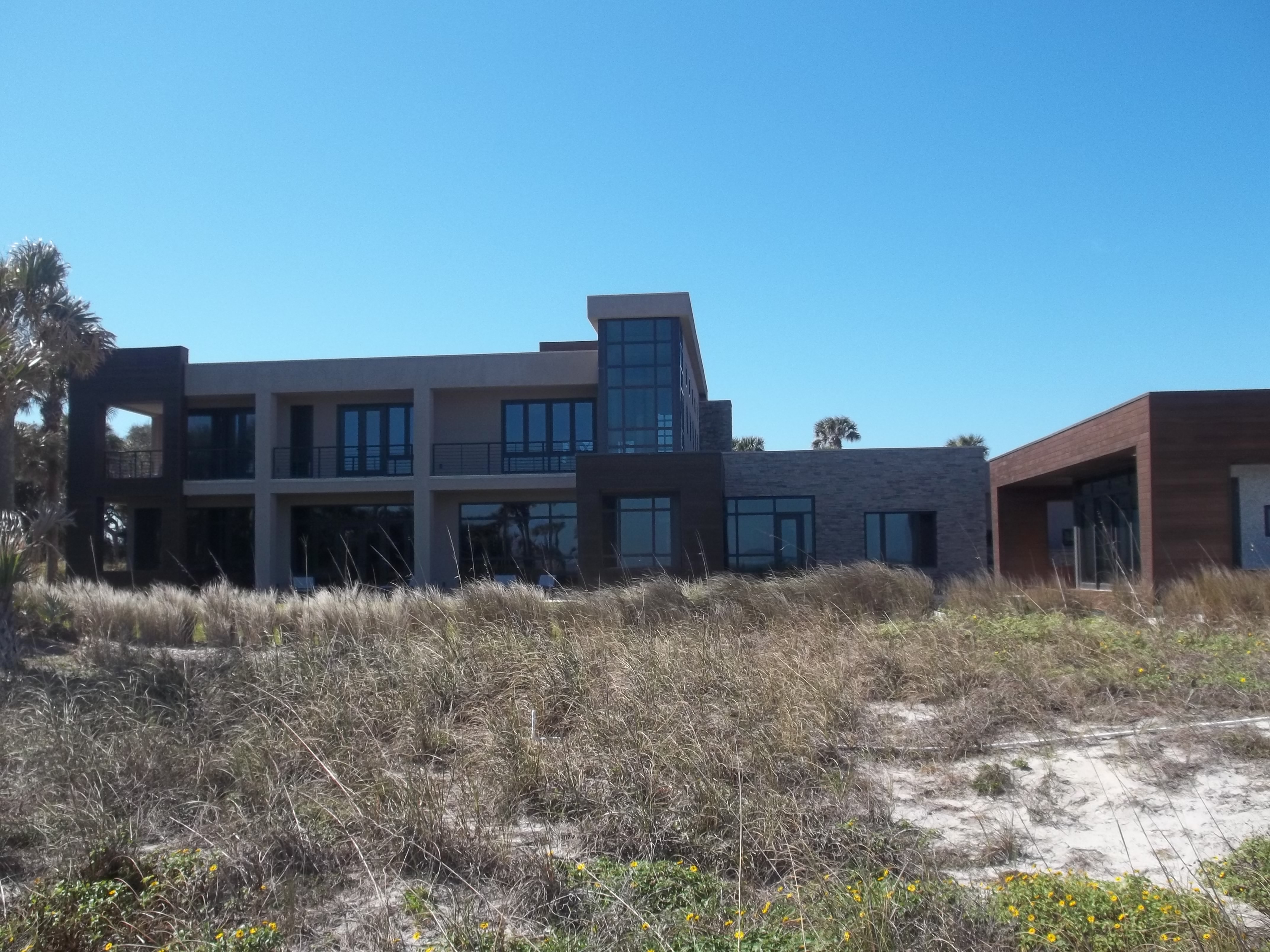 Modern two-story beachfront home with large windows and balcony behind coastal dunes.