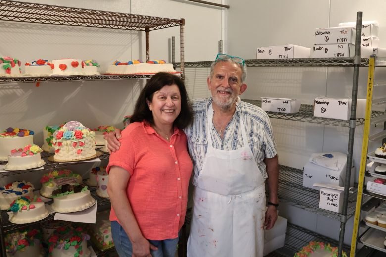 A man in a white apron and a woman in a coral top smiling together in a bakery storage room with decorated cakes on shelves behind them.