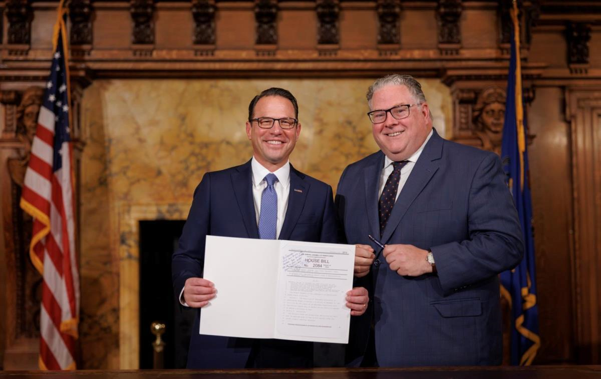 Two men in suits hold a signed document in an ornate government chamber with flags.