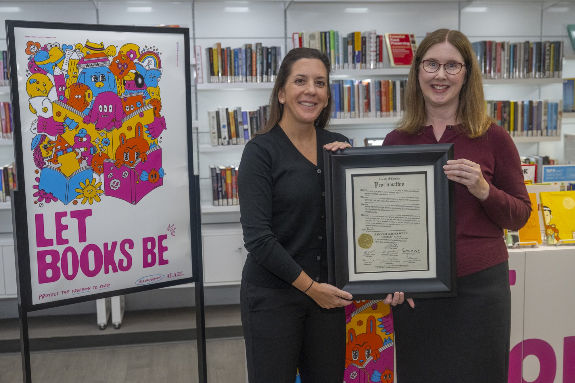Two women hold a framed proclamation beside a Banned Books Week poster in a library.