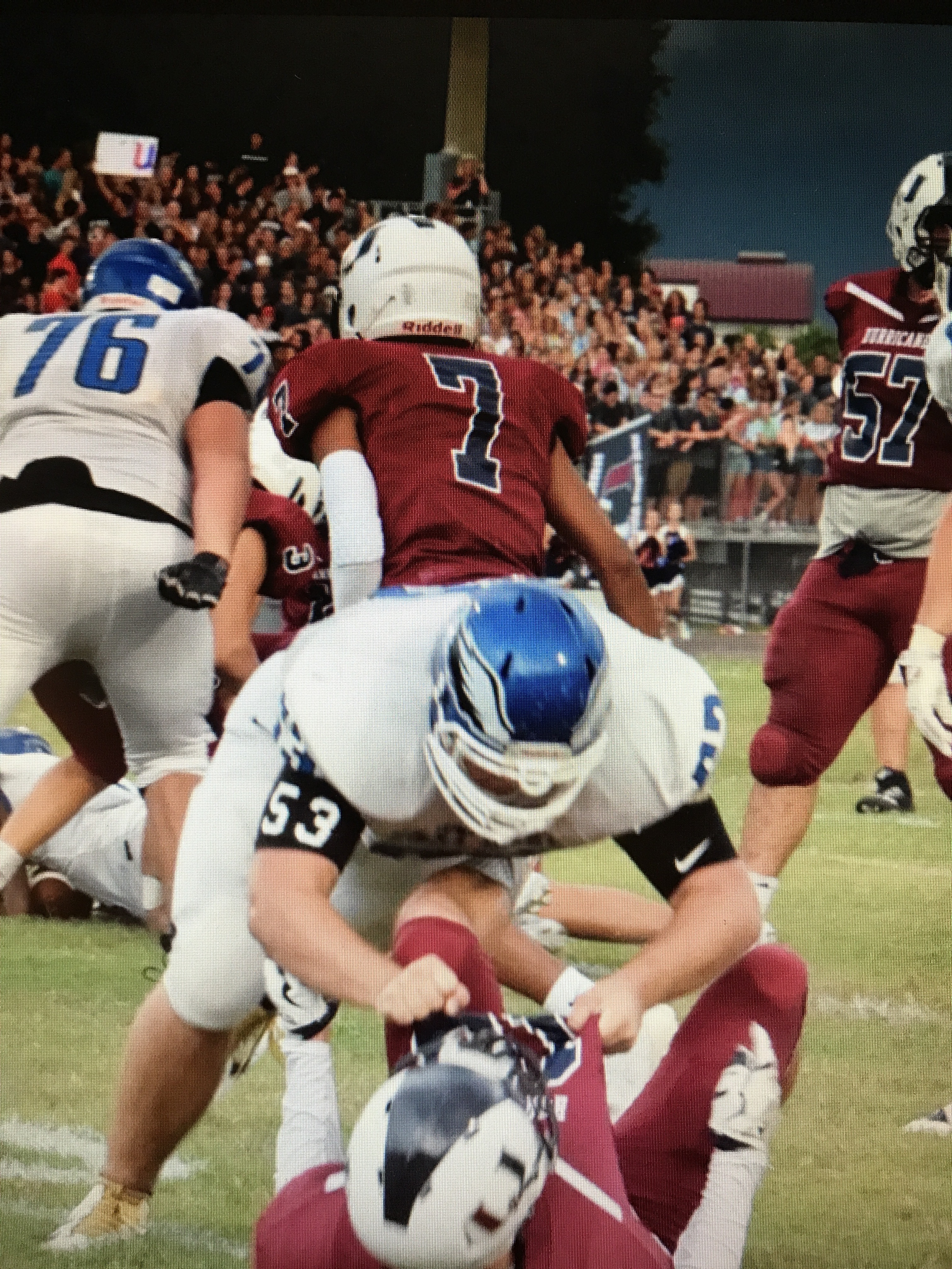 Football players in maroon and white uniforms collide near the line of scrimmage before a packed crowd.