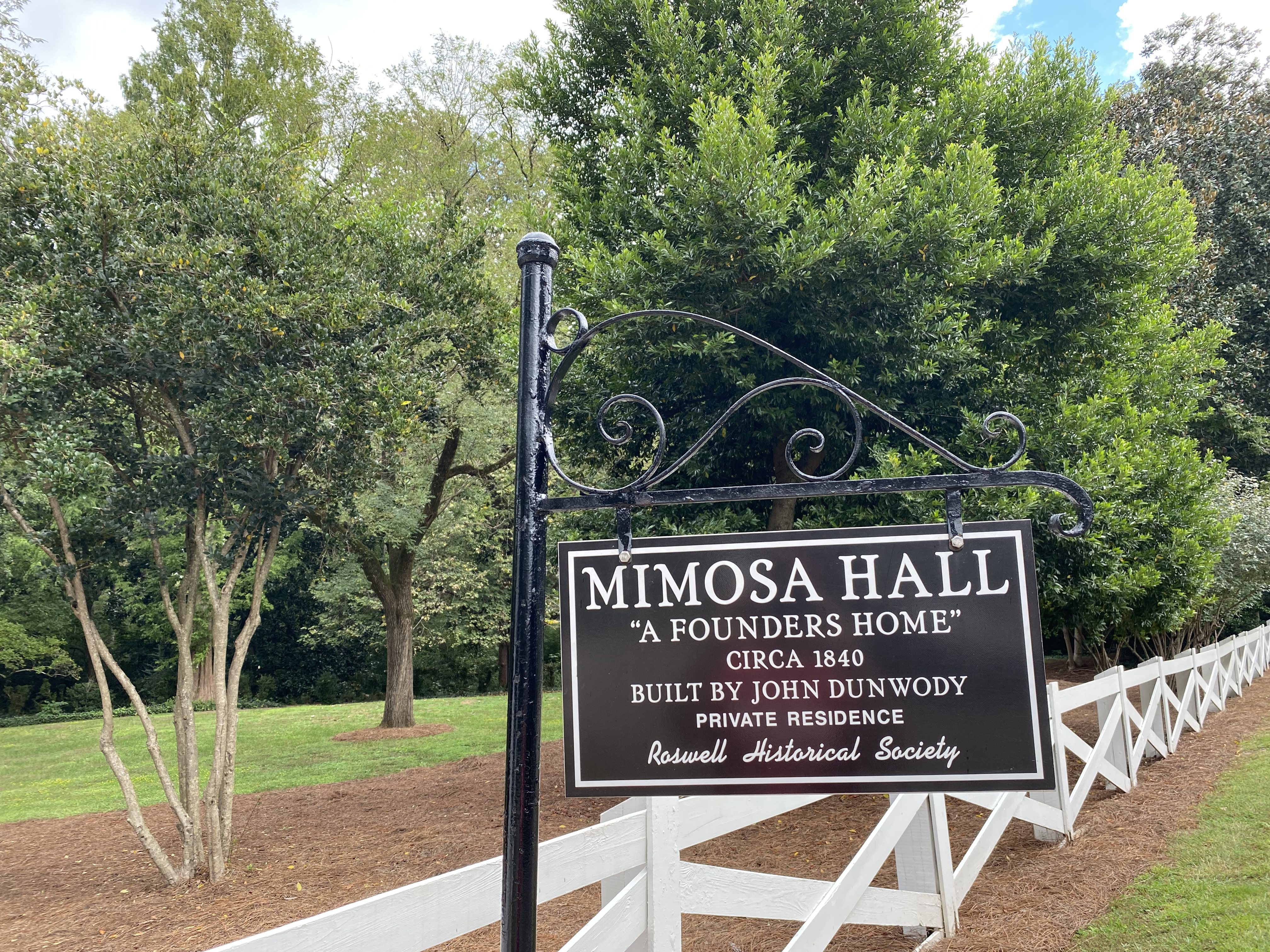 Black sign on a metal post reading 'Mimosa Hall – A Founders Home – Circa 1840' with a white fence and trees.