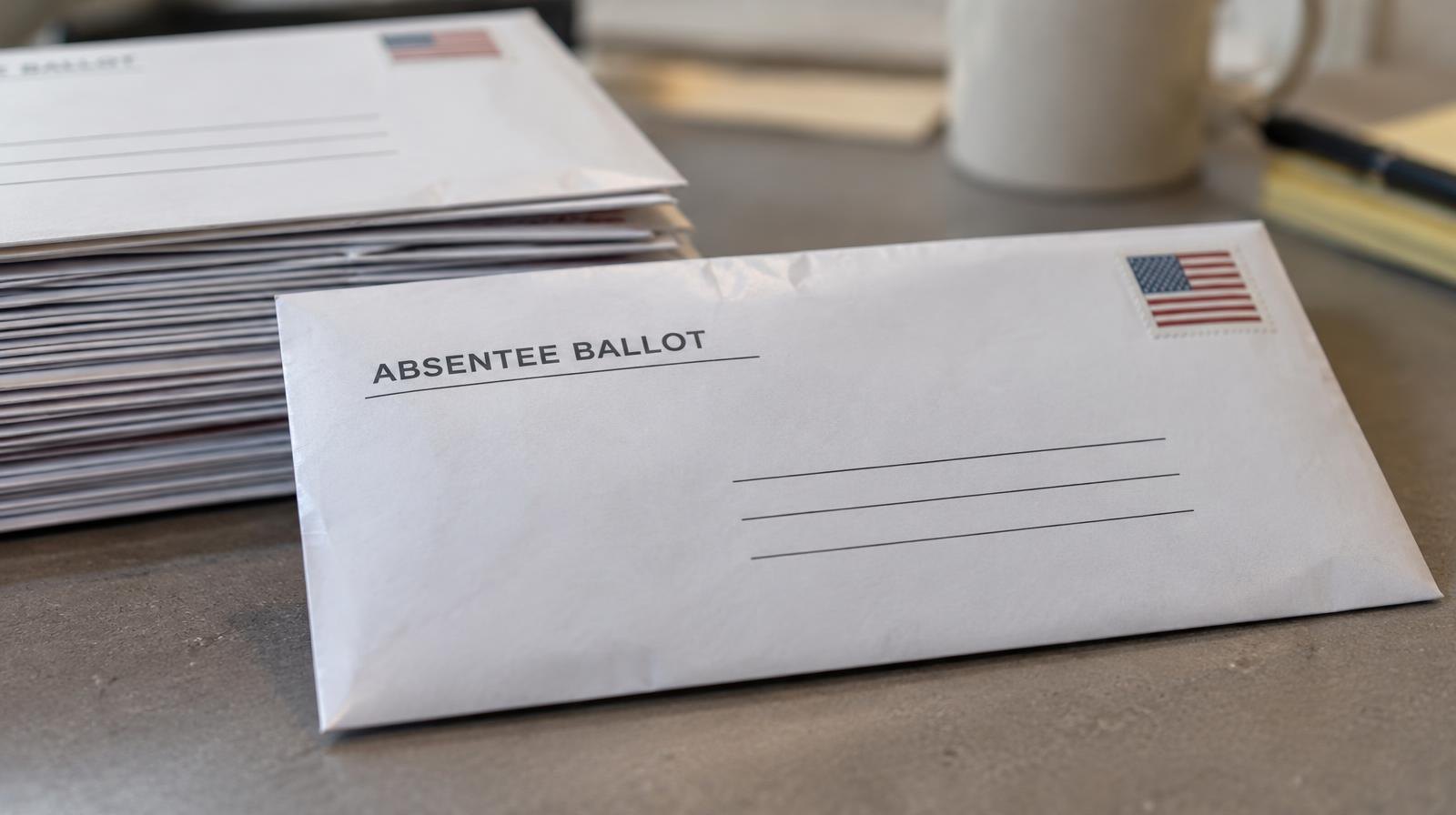 A sealed absentee ballot envelope resting beside a stack of similar envelopes on a flat surface.