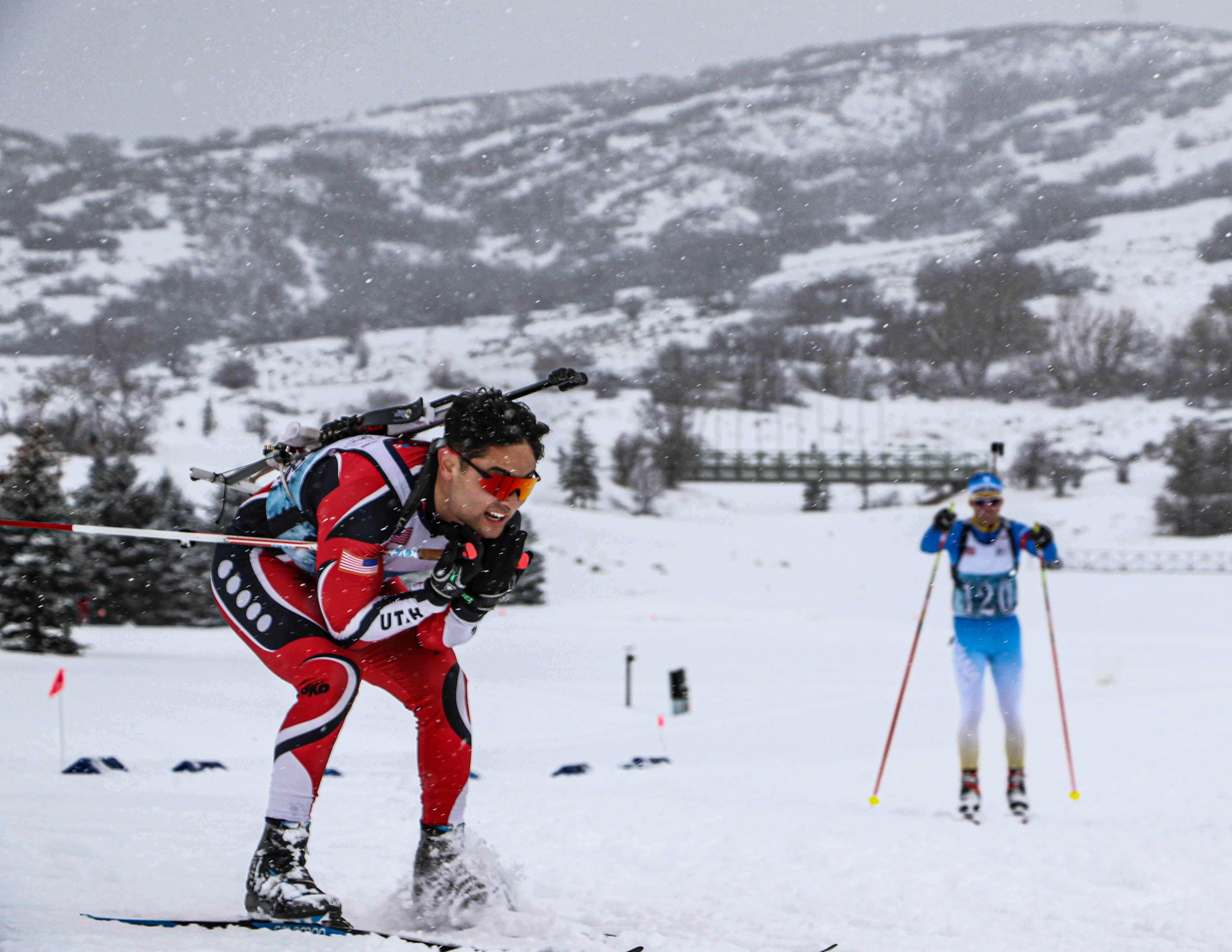 Two biathlon skiers with rifles on their backs race through falling snow.