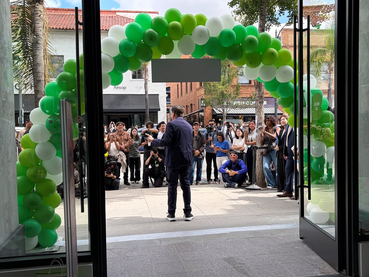 Crowd gathered outside a store entrance framed by a green and white balloon arch on a pedestrian retail street.