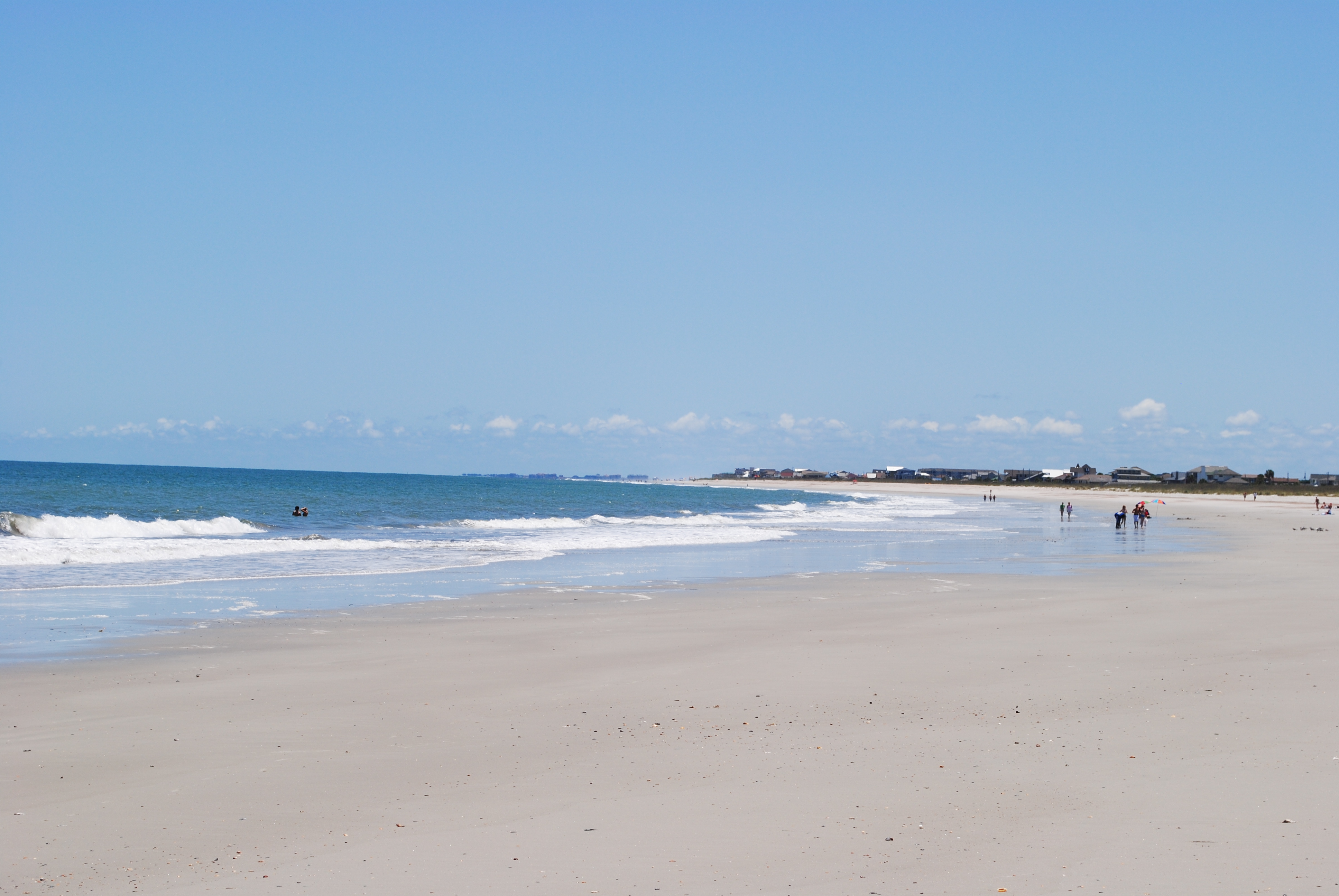 Wide sandy beach with gentle surf and a coastal town on the horizon.