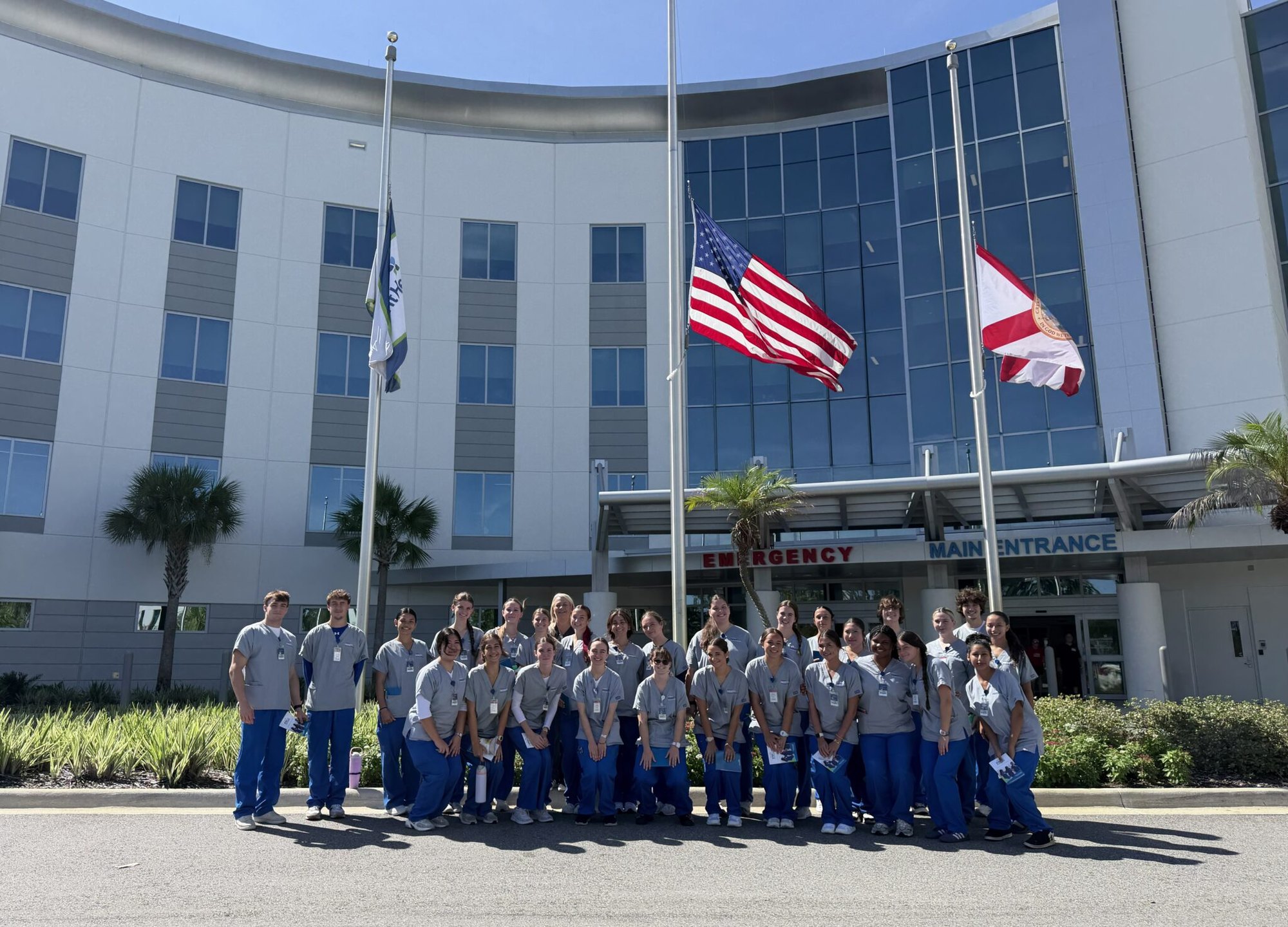 Group of students in scrubs posing outside a hospital emergency and main entrance.