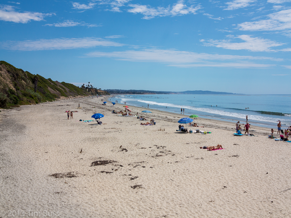 Sandy beach with scattered beachgoers and coastal bluffs under a partly cloudy sky.