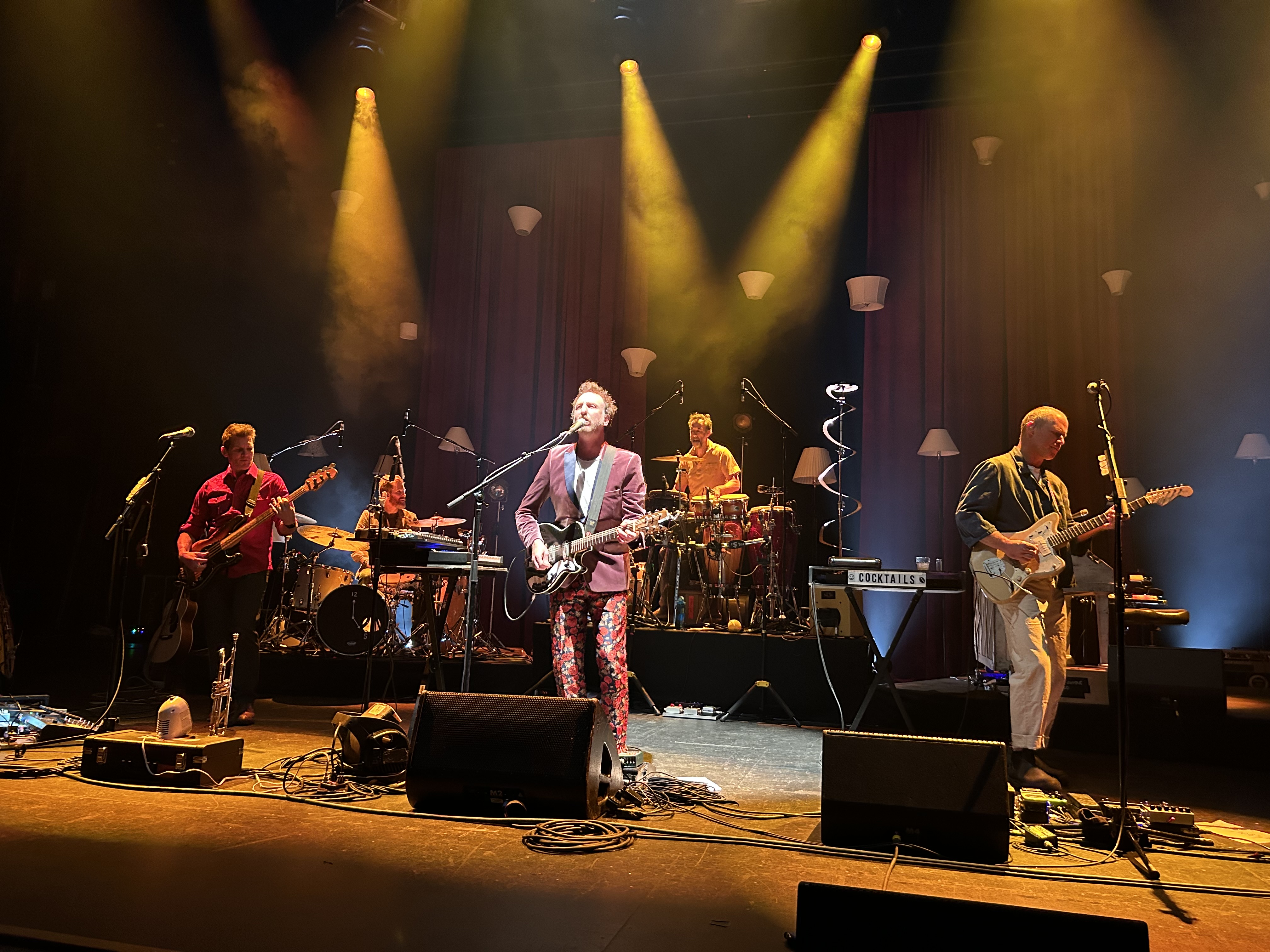Band performing on a brightly lit stage with yellow and green spotlights at a concert venue.
