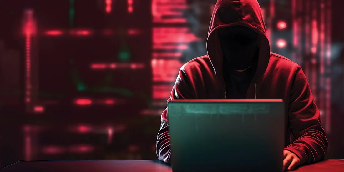 Hooded figure at a laptop in a darkened room with red digital background lighting.