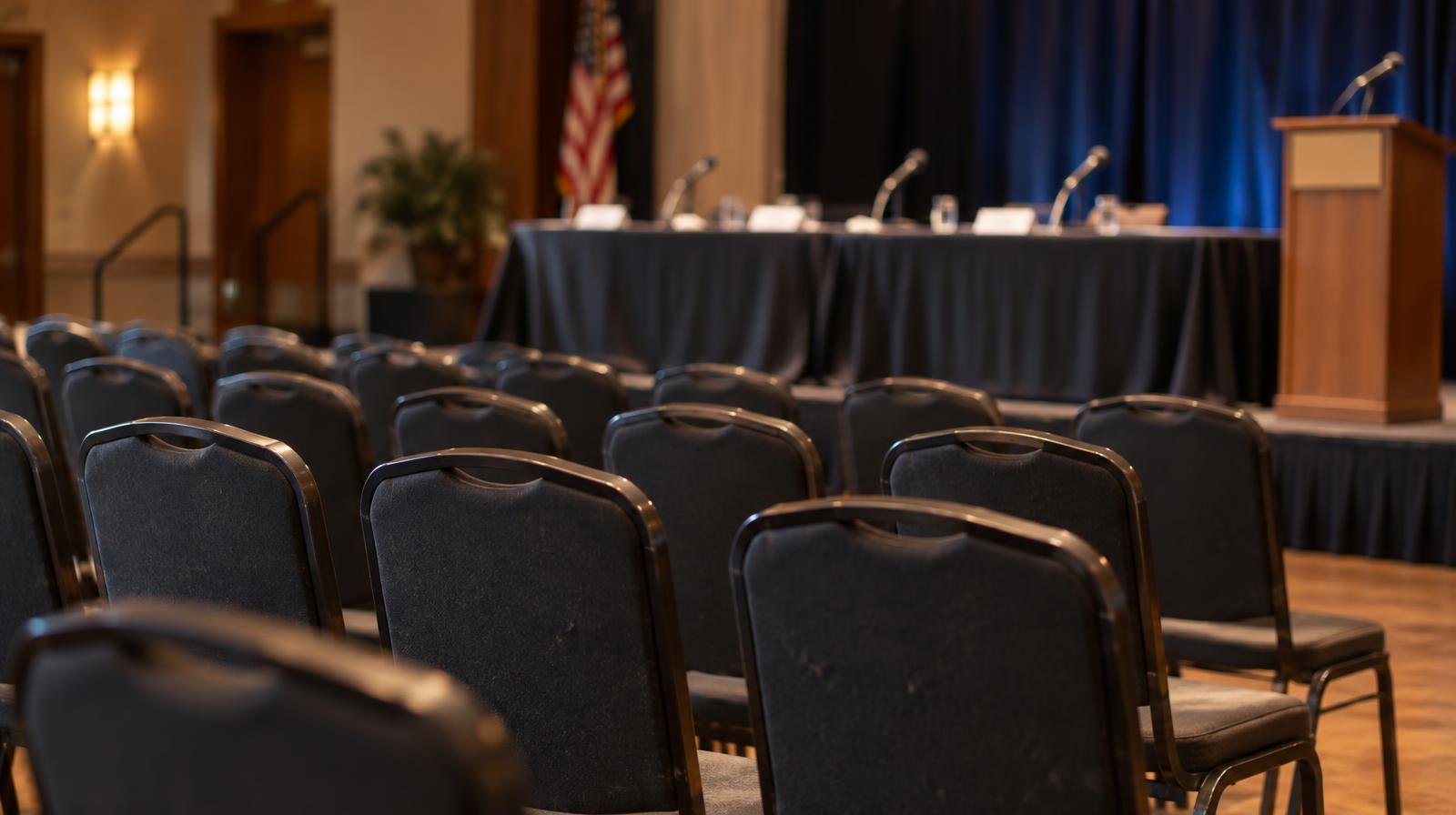 Empty rows of chairs facing a table with microphones in a meeting room.