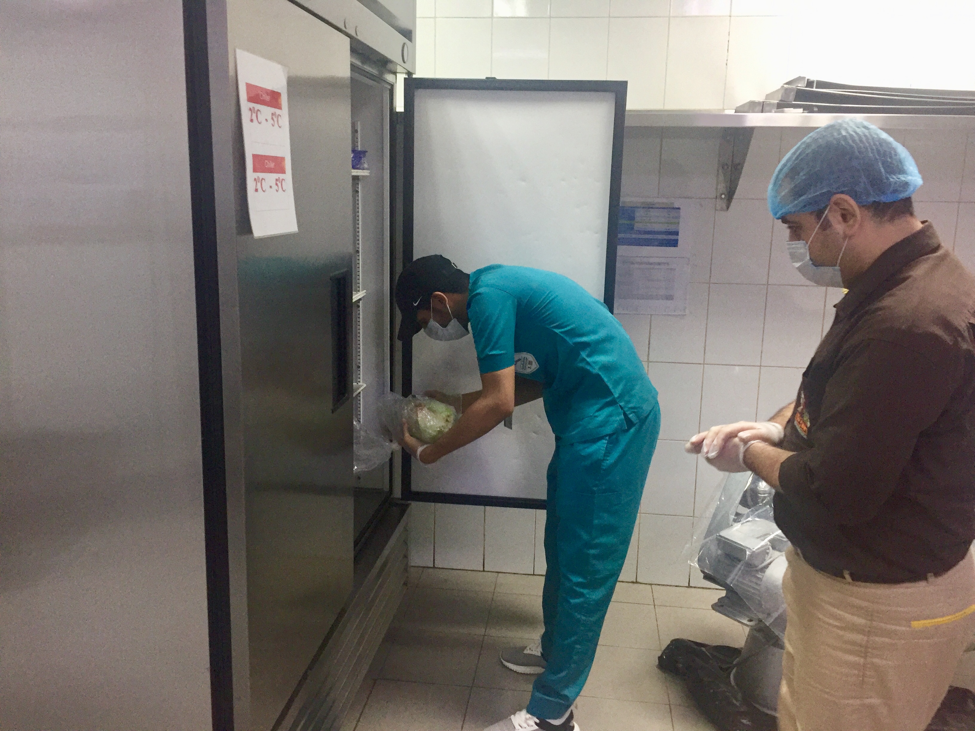 Two workers in protective gear inspect an open stainless steel refrigerator in a commercial kitchen.