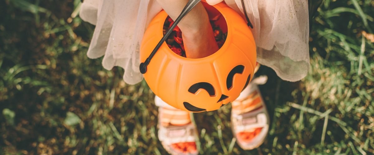 Child in costume holds an orange jack-o'-lantern shaped Halloween candy bucket.