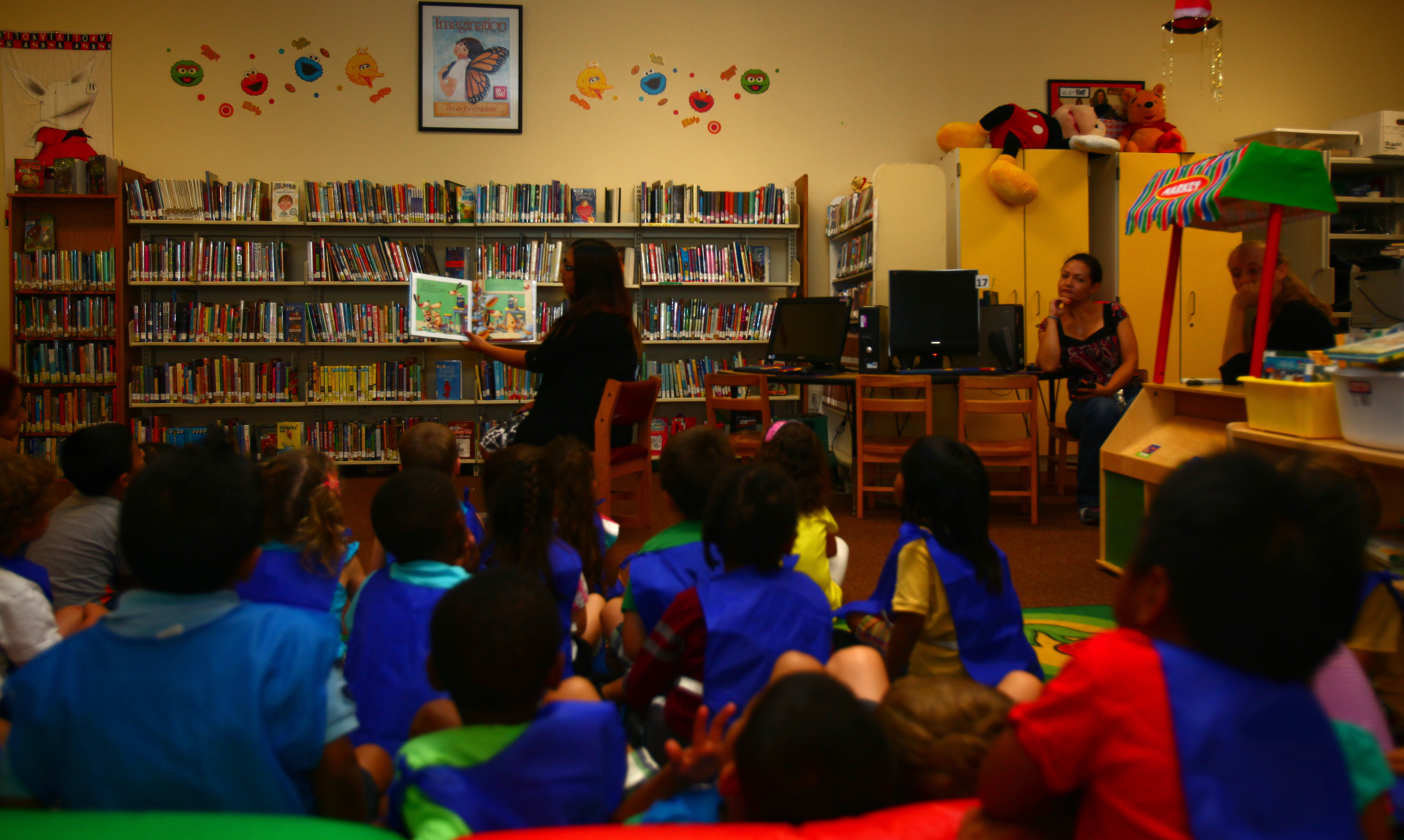 Children in colorful smocks sit on a library floor during a storytime session.