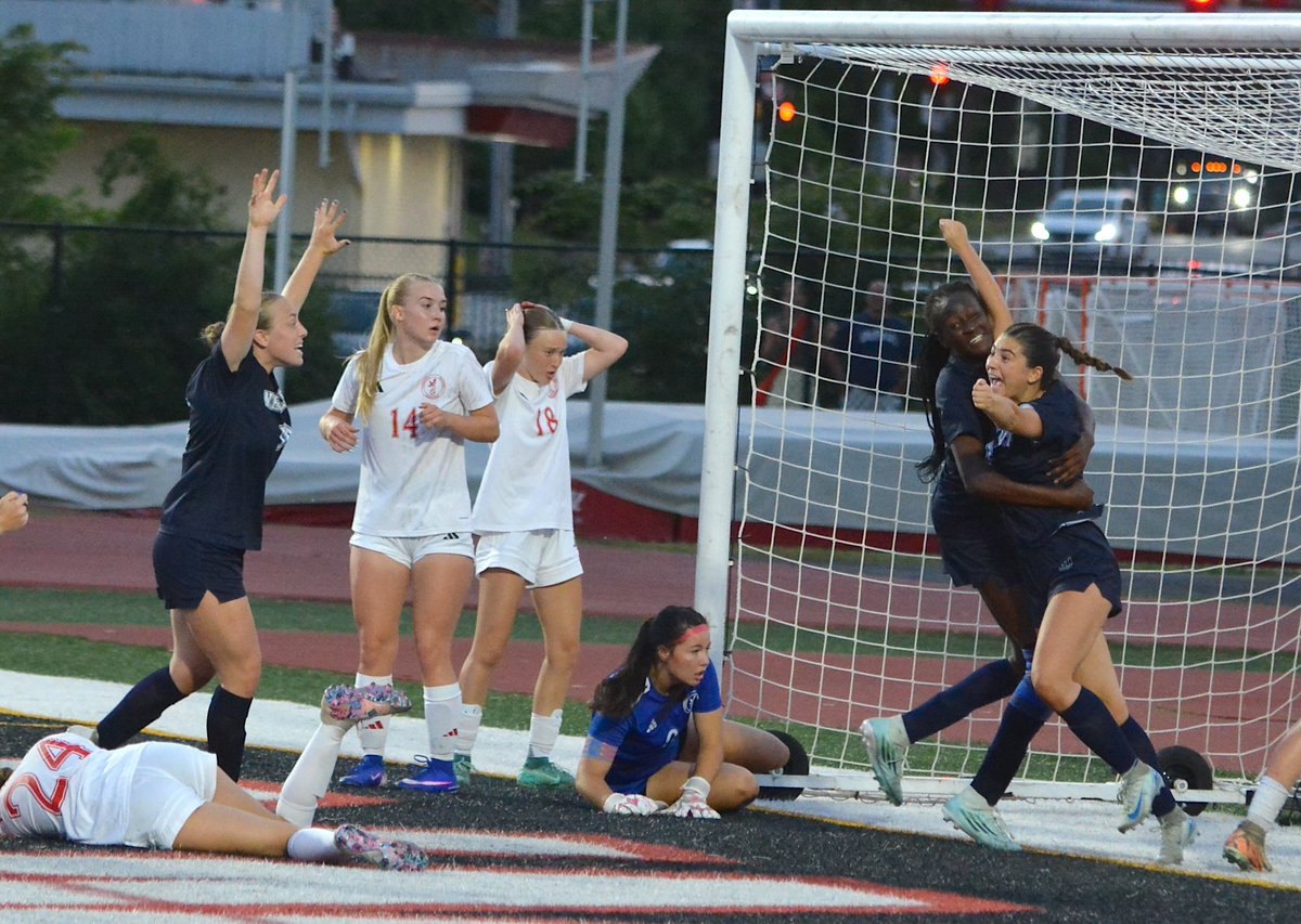 Girls soccer players react near the goal as a score is made, with some celebrating and others showing dismay.