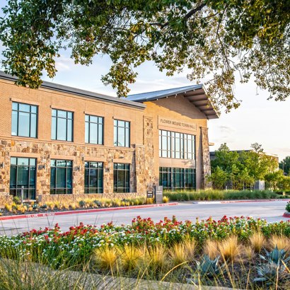 Two-story brick and stone municipal building with flower beds and oak trees at golden hour.