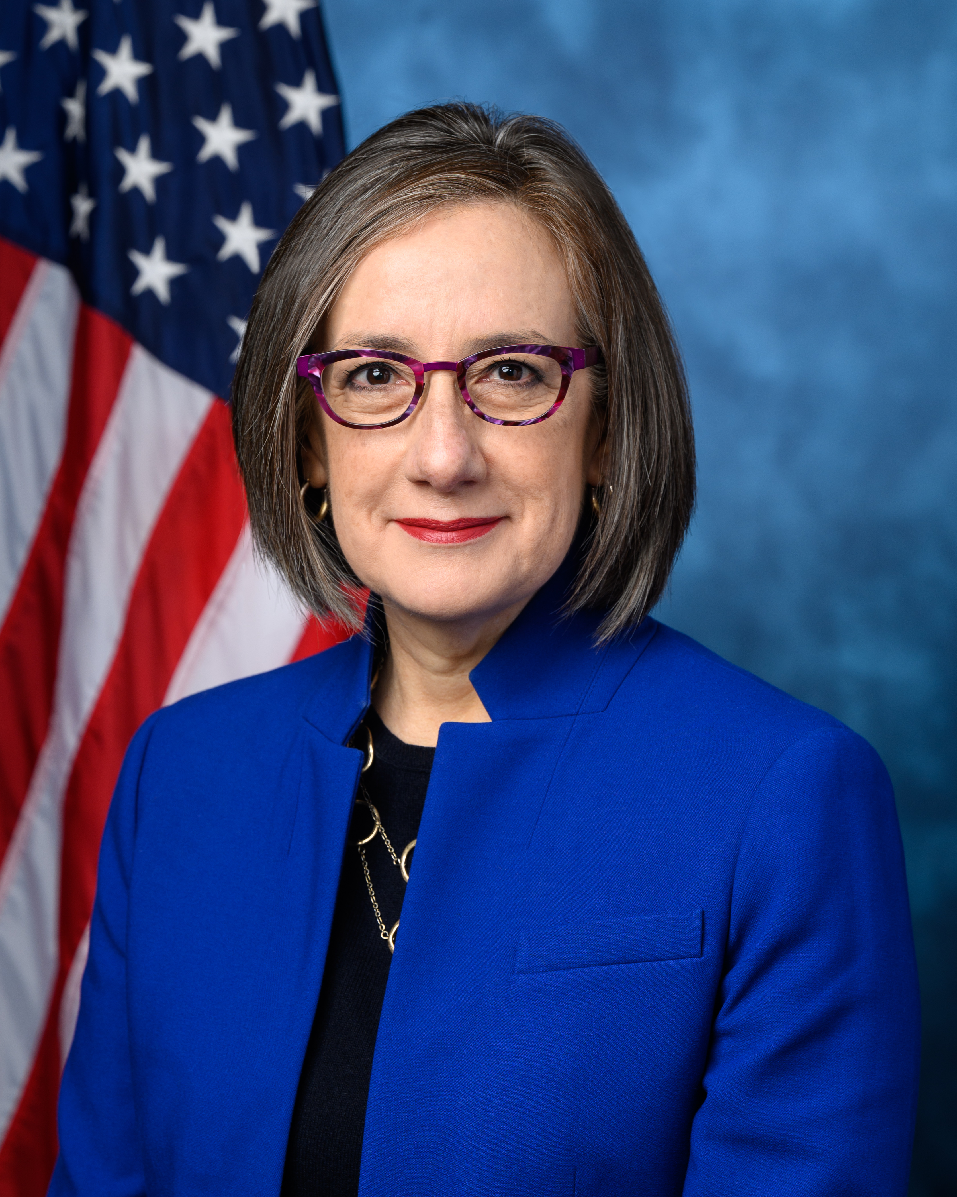 Woman in blue blazer and purple glasses posed before an American flag in a formal portrait.