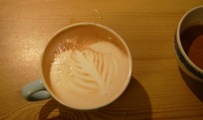 A cappuccino with leaf latte art in a white ceramic mug on a wooden table.