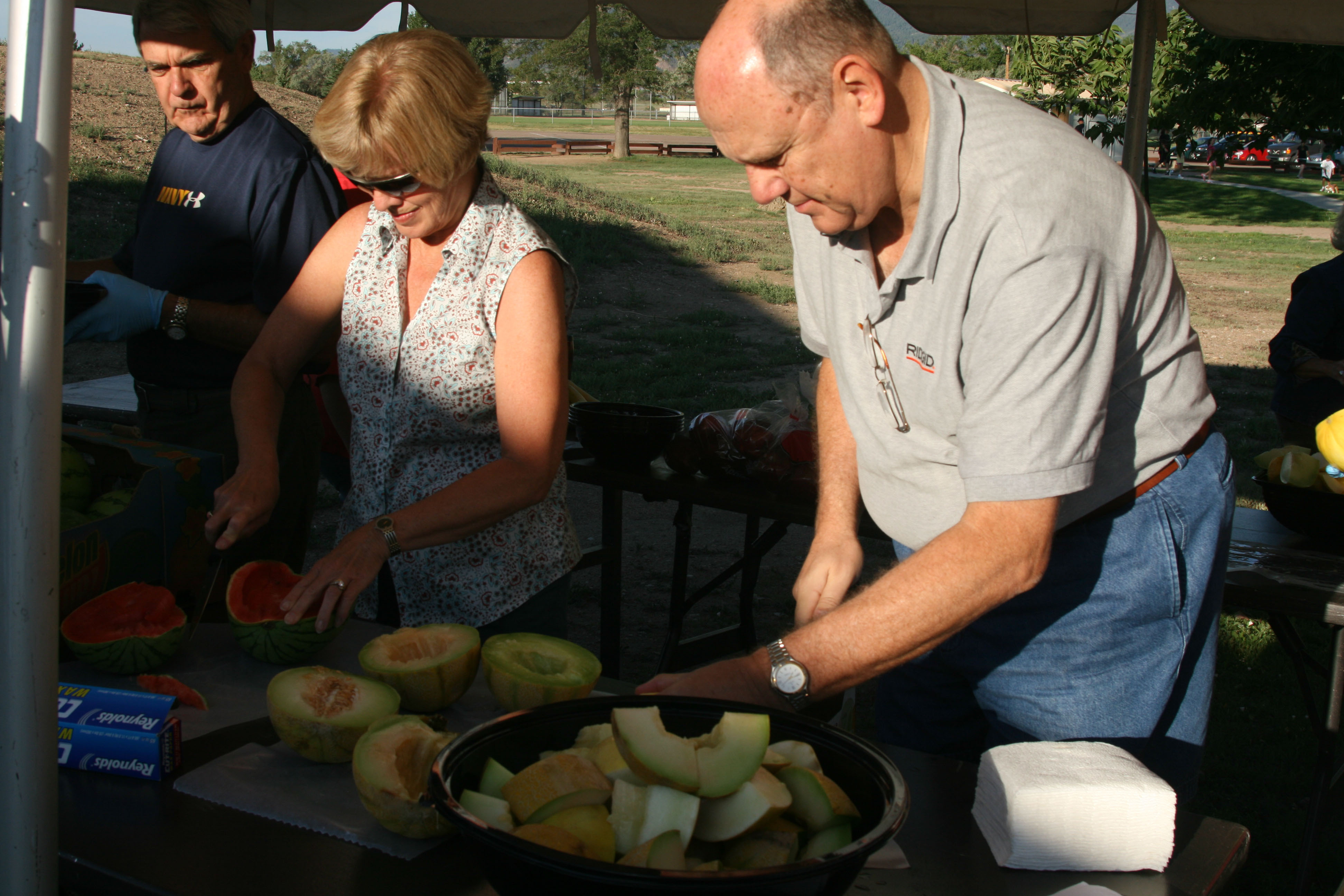 Two people cutting watermelon and cantaloupe at an outdoor table under a canopy.